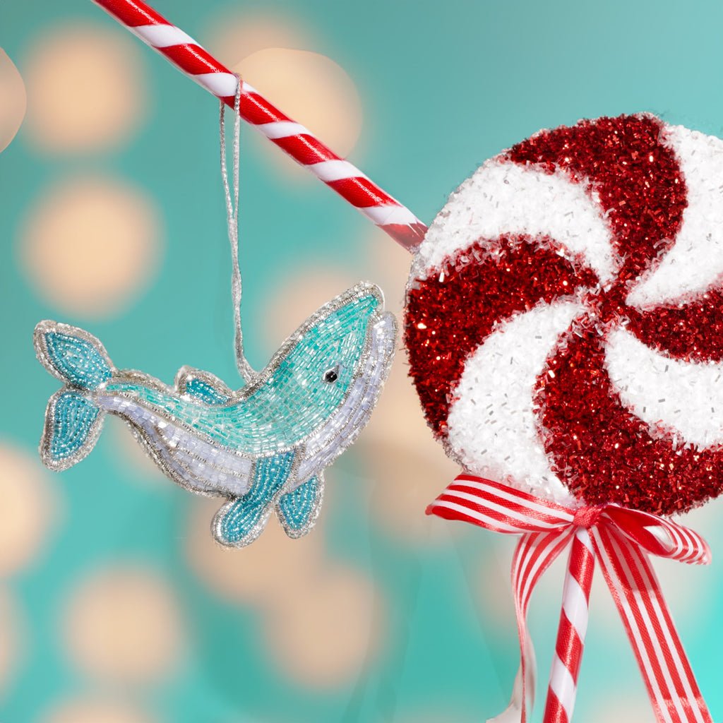 A turquoise and white beaded whale decoration with silver trim hanging beside a large red and white glittery peppermint lollipop tied with a red striped ribbon against a teal background with soft glowing lights.