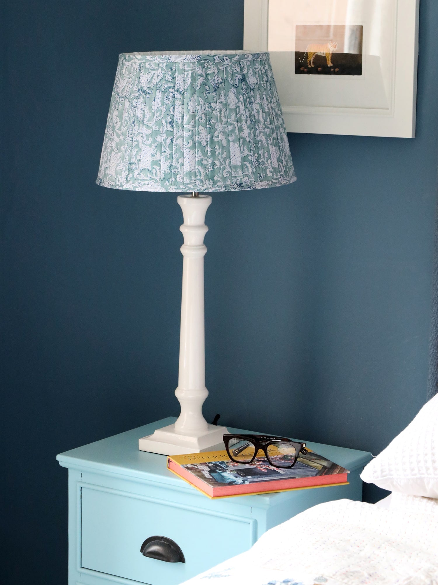 White Lacquer Finished lampbase with a pleated lampshade on a sideboard.There is a book and a pair of glasses on the side,on the wall behind is a picture.