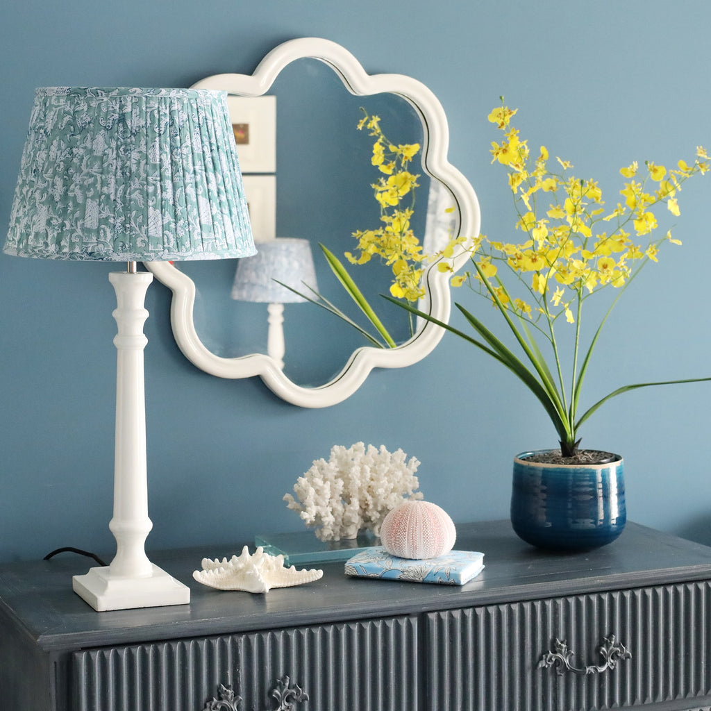 White Lacquer Godophin lampbase with a pleated lampshade on a sideboard.On the sideboard are several shells, flowers in a pot and a small book.Behind on the wall is a wave mirror