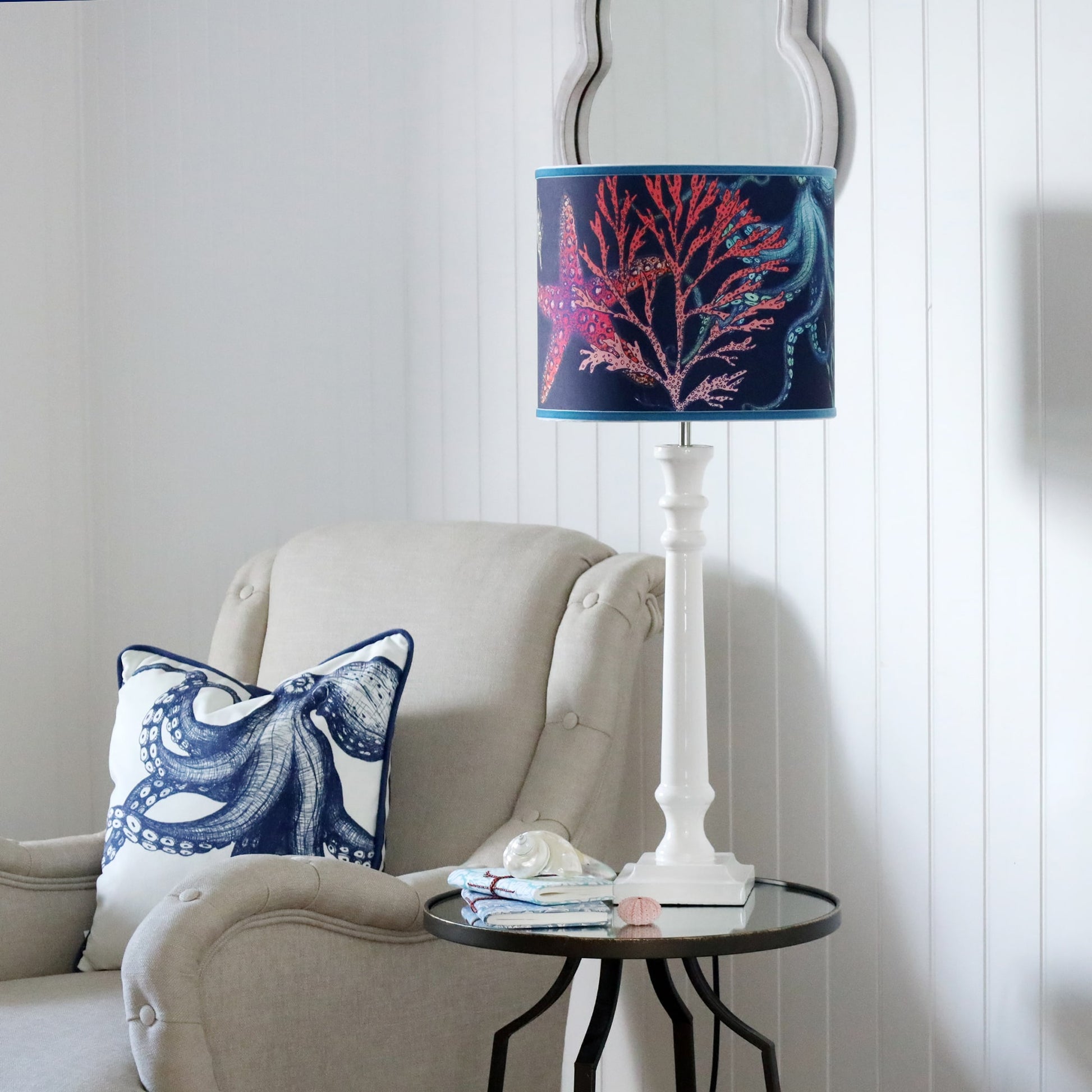White Lacquer Godolphin lamp base with an Reef lampshade on a mirror table.On the table is a couple of books with a shell placed on it.Next to the table is a chair with an octopus cushion.Behind on the wall is a wave mirror.