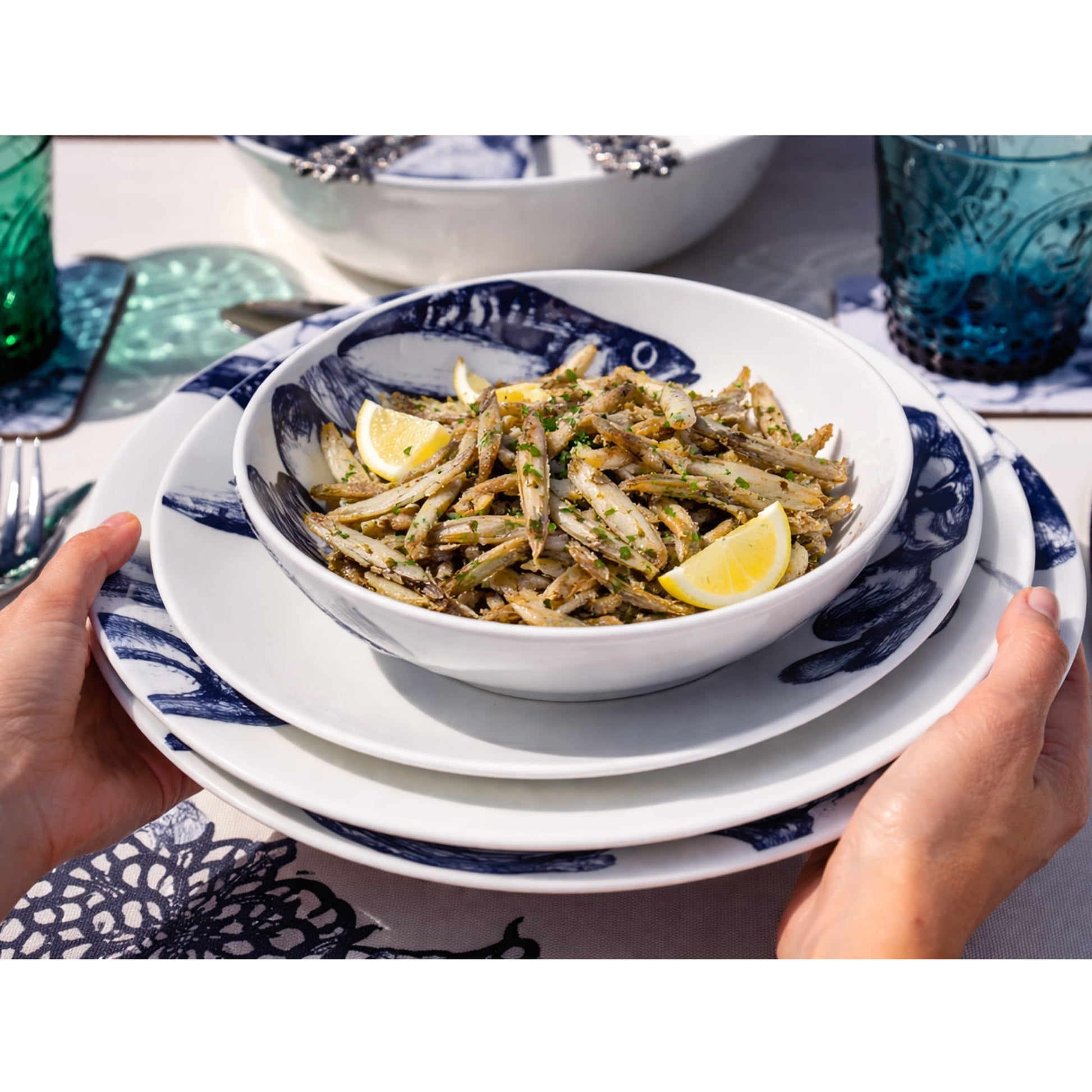 Bowl of salad with lemon wedges on a stack of plates with mackerel design held by hands.