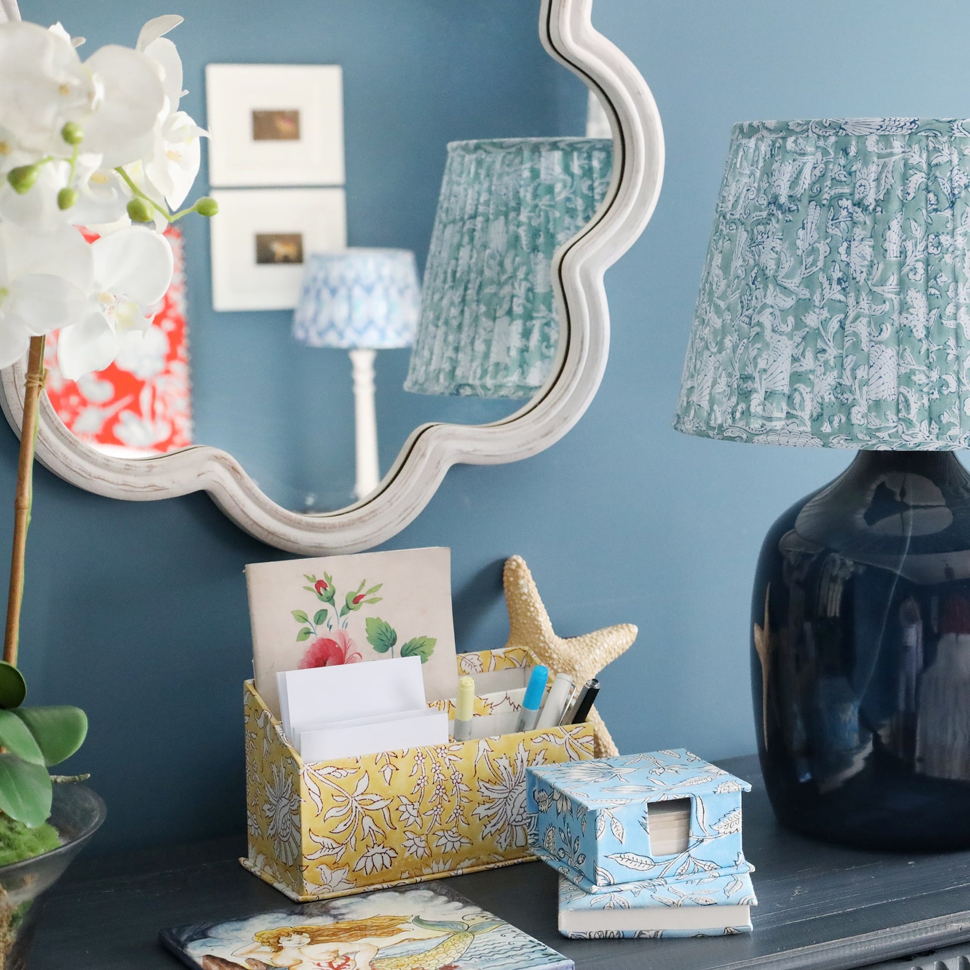 Whitewashed wooden wavey edged shaped mirror on the wall.In front is a sideboard with an orchid plant,some stationery  and one of our navy lamp bases and a pleated lampshade