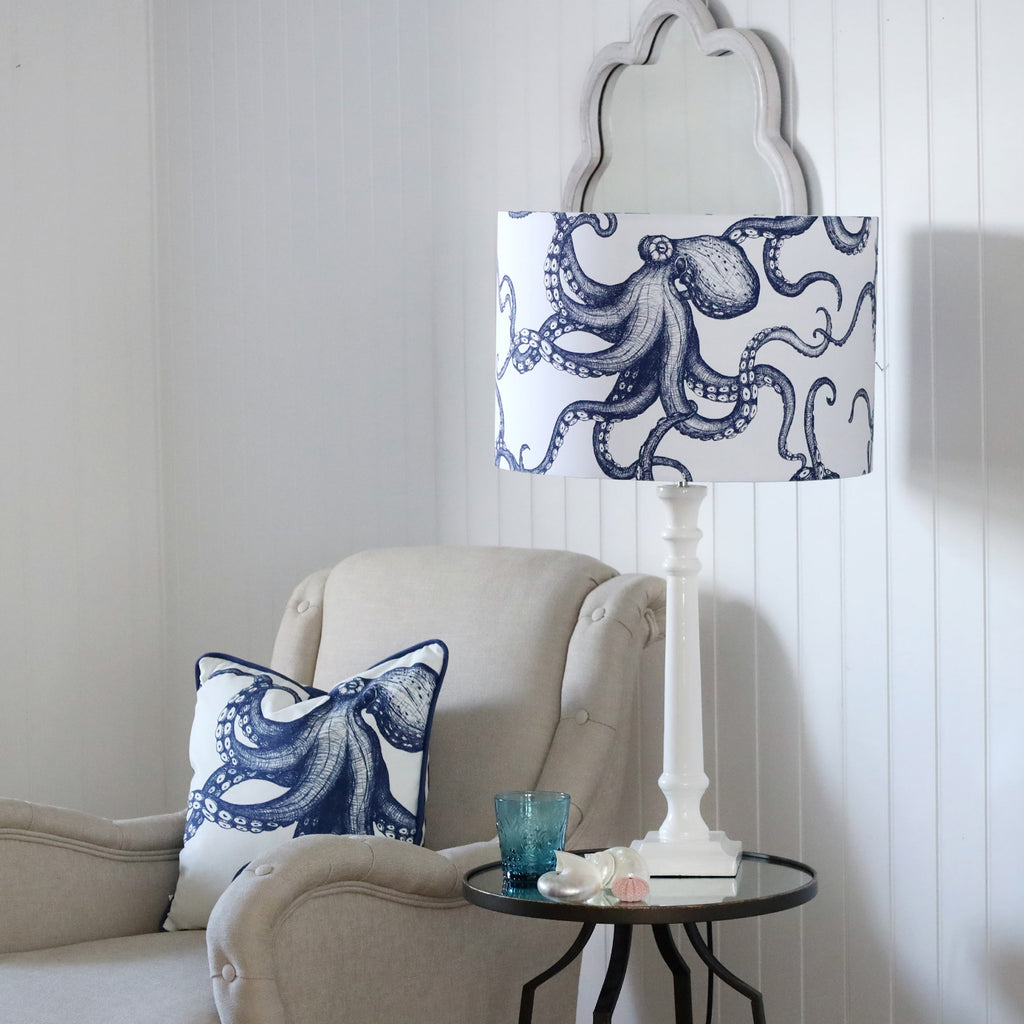 White Lacquer Godolphin lamp base with an octopus lampshade on a mirror table.On the table is a glass with some shells placed on it.Next to the table is a chair with an octopus cushion.Behind on the wall is a wave mirror.