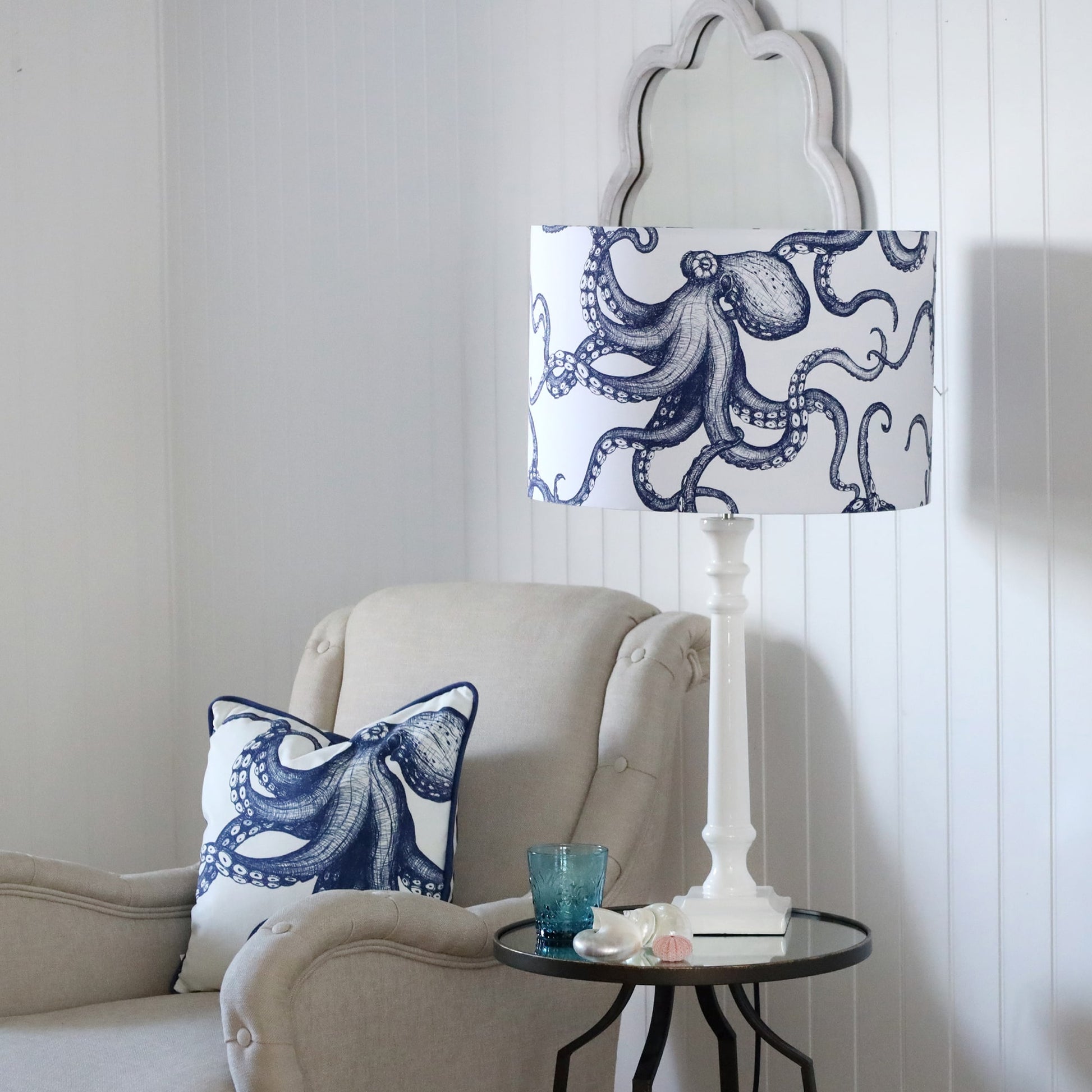 White Lacquer Godolphin lamp base with an octopus lampshade on a mirror table.On the table is a glass with some shells placed on it.Next to the table is a chair with an octopus cushion.Behind on the wall is a wave mirror.