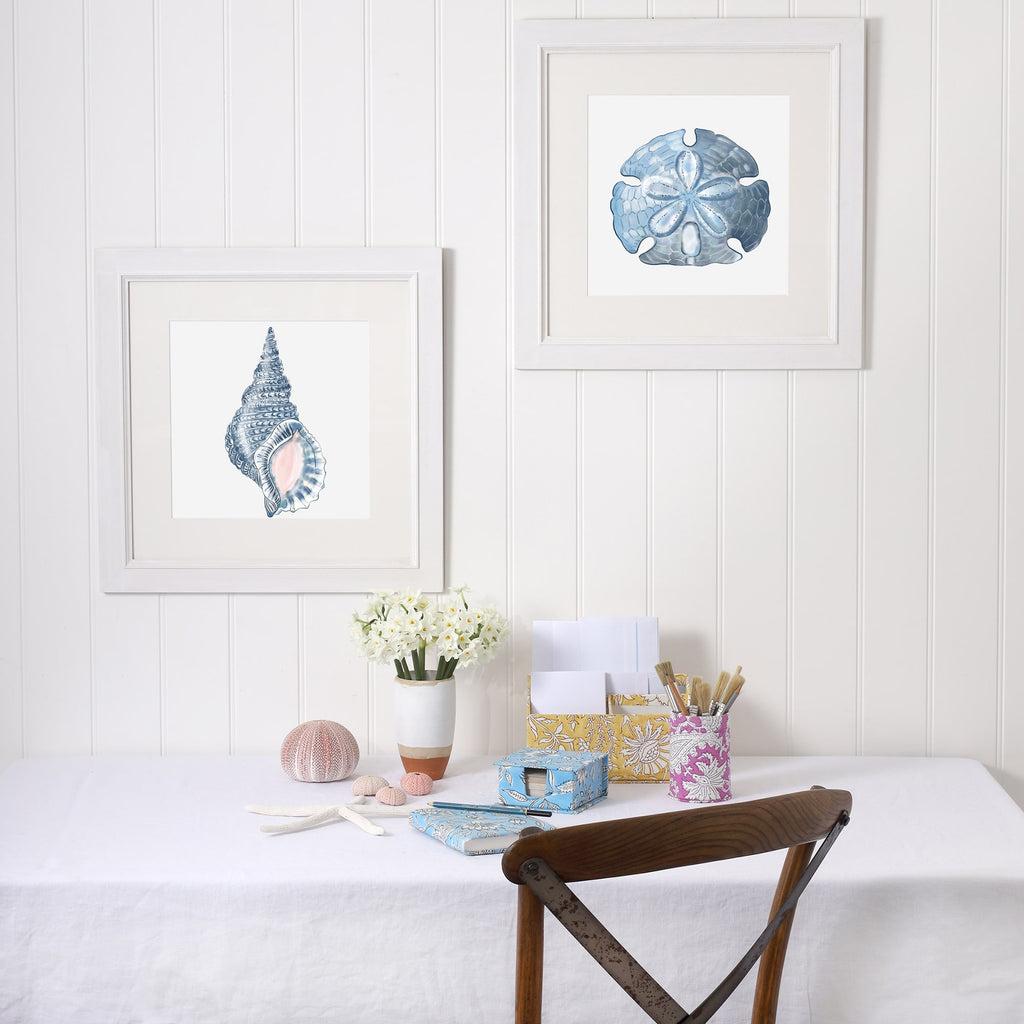 A serene workspace with a coastal theme, featuring two framed marine-inspired artworks on a white paneled wall. One artwork showcases a blue sand dollar, while the other features a detailed whelk shell. Below, a white linen-covered table is adorned with decorative stationery in floral prints, a vase of white flowers, a pink sea urchin, and seashells. A wooden chair with a curved metal frame adds a rustic touch to the calming, ocean-inspired setting.