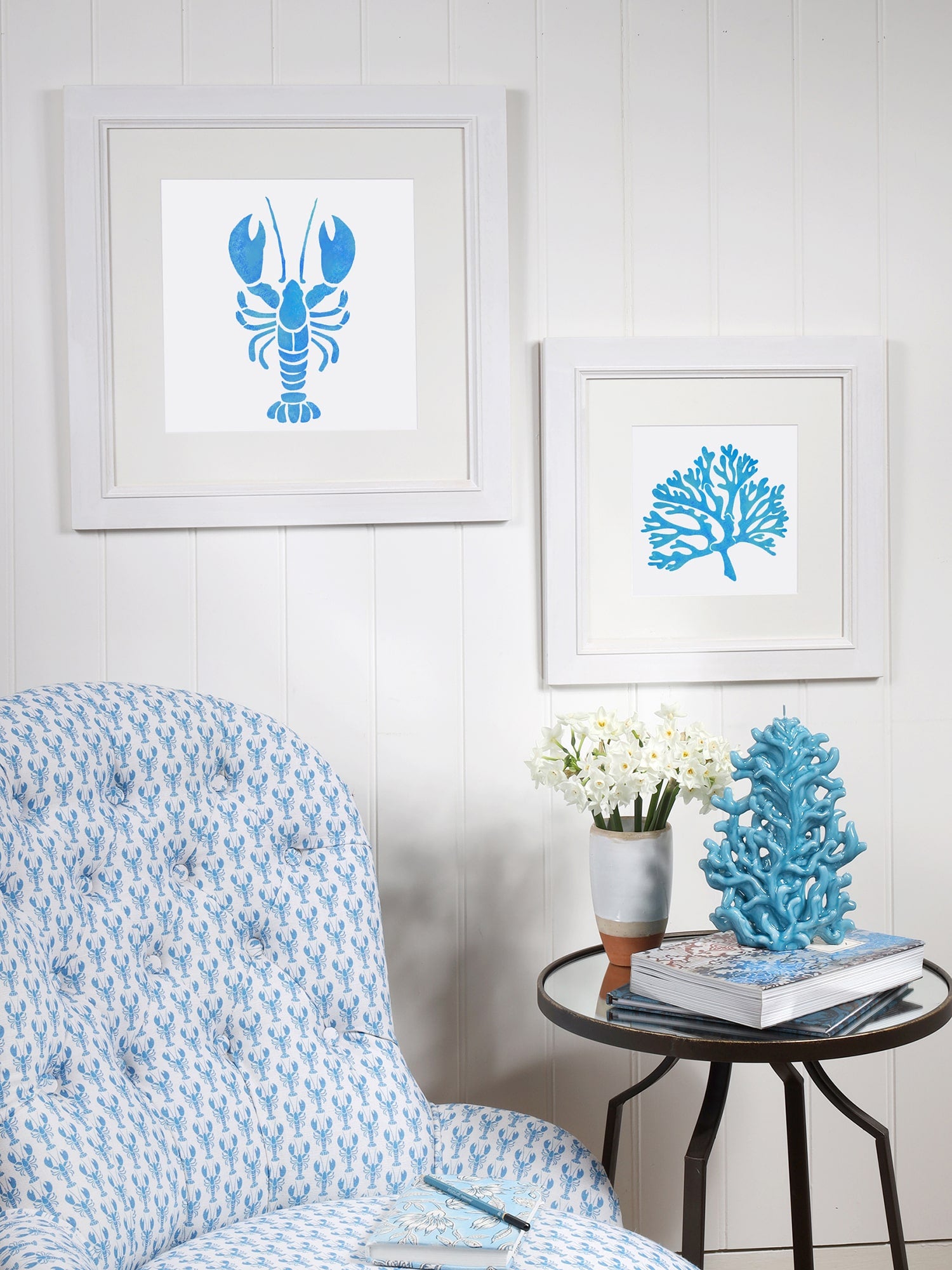 A coastal-inspired interior setting featuring two framed blue stencil-style illustrations of a lobster and coral, mounted on a white paneled wall. Below, a blue and white lobster-patterned upholstered chair is positioned beside a round metal side table. The table is decorated with a blue ceramic coral sculpture, a vase of white flowers, and a stack of books. A light blue notebook and pen rest on the chair's cushion, complementing the nautical theme.