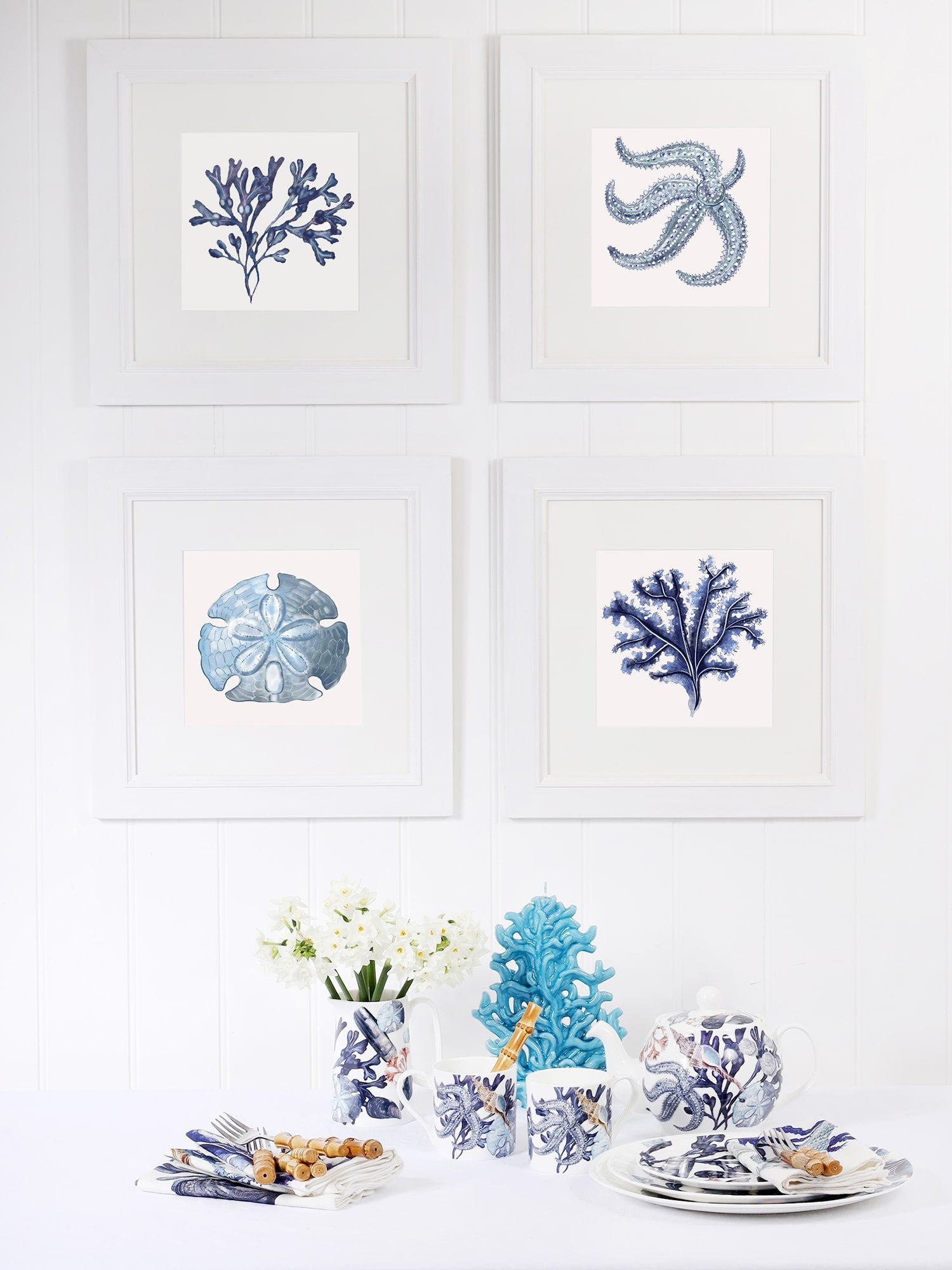  A dining setup with four framed coastal illustrations on a white wall, featuring seaweed, a starfish, a sand dollar, and coral. Below, a table is set with blue and white marine-themed crockery, napkins, and wooden utensils, complemented by white flowers and a decorative blue coral ornament.