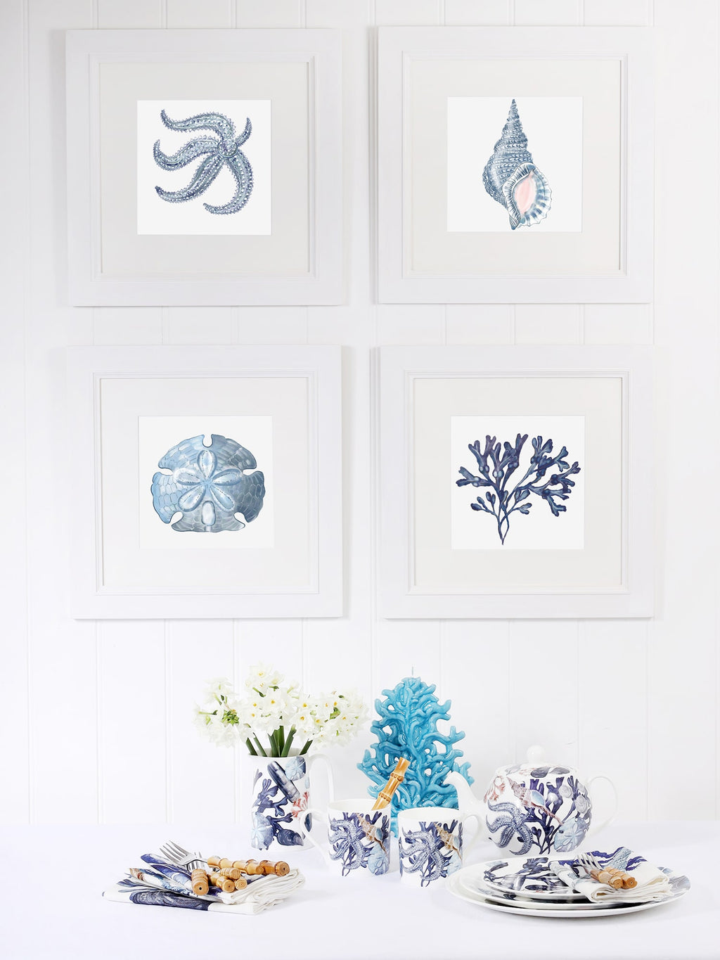 A coastal-themed dining setup with four framed marine-inspired artworks in white frames on a white paneled wall. The artworks feature illustrations of a blue starfish, a whelk shell, a sand dollar, and coral. Below, the table is set with ocean-themed tableware, including mugs, plates, and a teapot adorned with blue and white sea life illustrations. A vase with white flowers, a decorative blue coral sculpture, and neatly arranged napkins with cutlery complete the serene, nautical-inspired aesthetic.