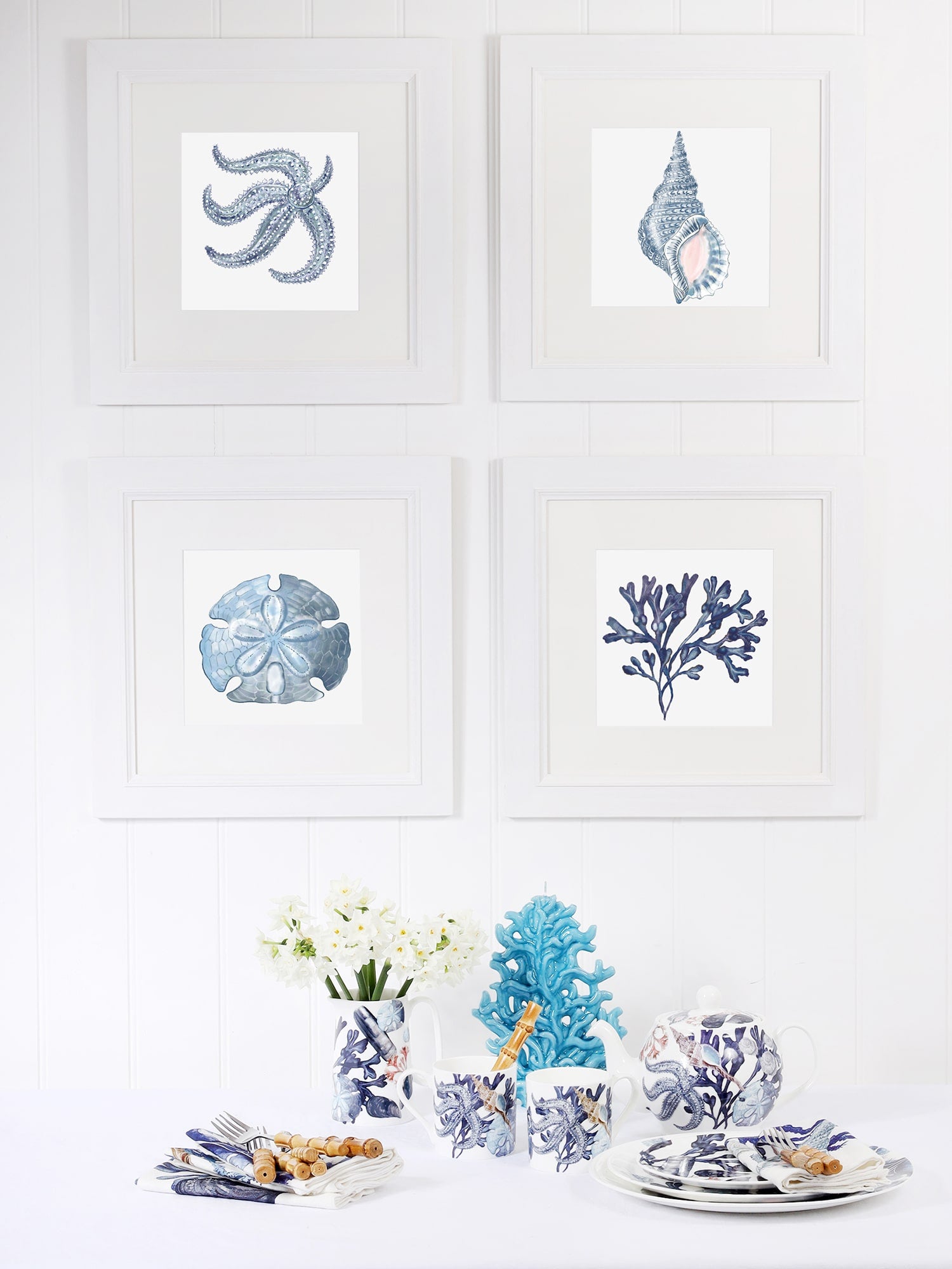 A coastal-themed dining setup with four framed marine-inspired artworks in white frames on a white paneled wall. The artworks feature illustrations of a blue starfish, a whelk shell, a sand dollar, and coral. Below, the table is set with ocean-themed tableware, including mugs, plates, and a teapot adorned with blue and white sea life illustrations. A vase with white flowers, a decorative blue coral sculpture, and neatly arranged napkins with cutlery complete the serene, nautical-inspired aesthetic.