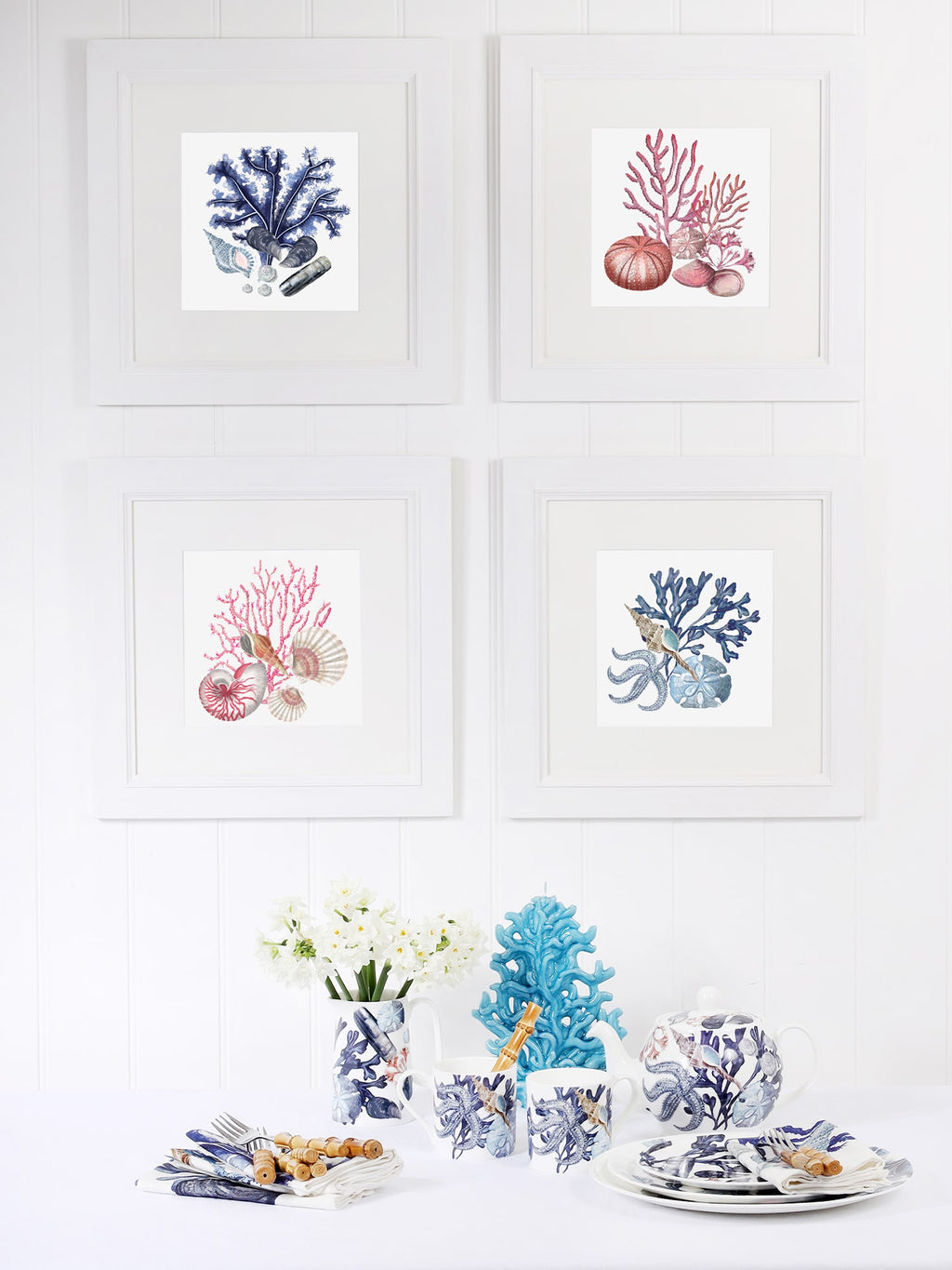 A coastal-themed dining setup with four framed coral and seashell prints in white frames on a white paneled wall. Below, a table is set with marine-inspired tableware, including mugs, plates, and a teapot featuring blue and white sea life illustrations. A vase with white flowers, a blue coral sculpture, and neatly arranged napkins with cutlery complete the ocean-inspired aesthetic.