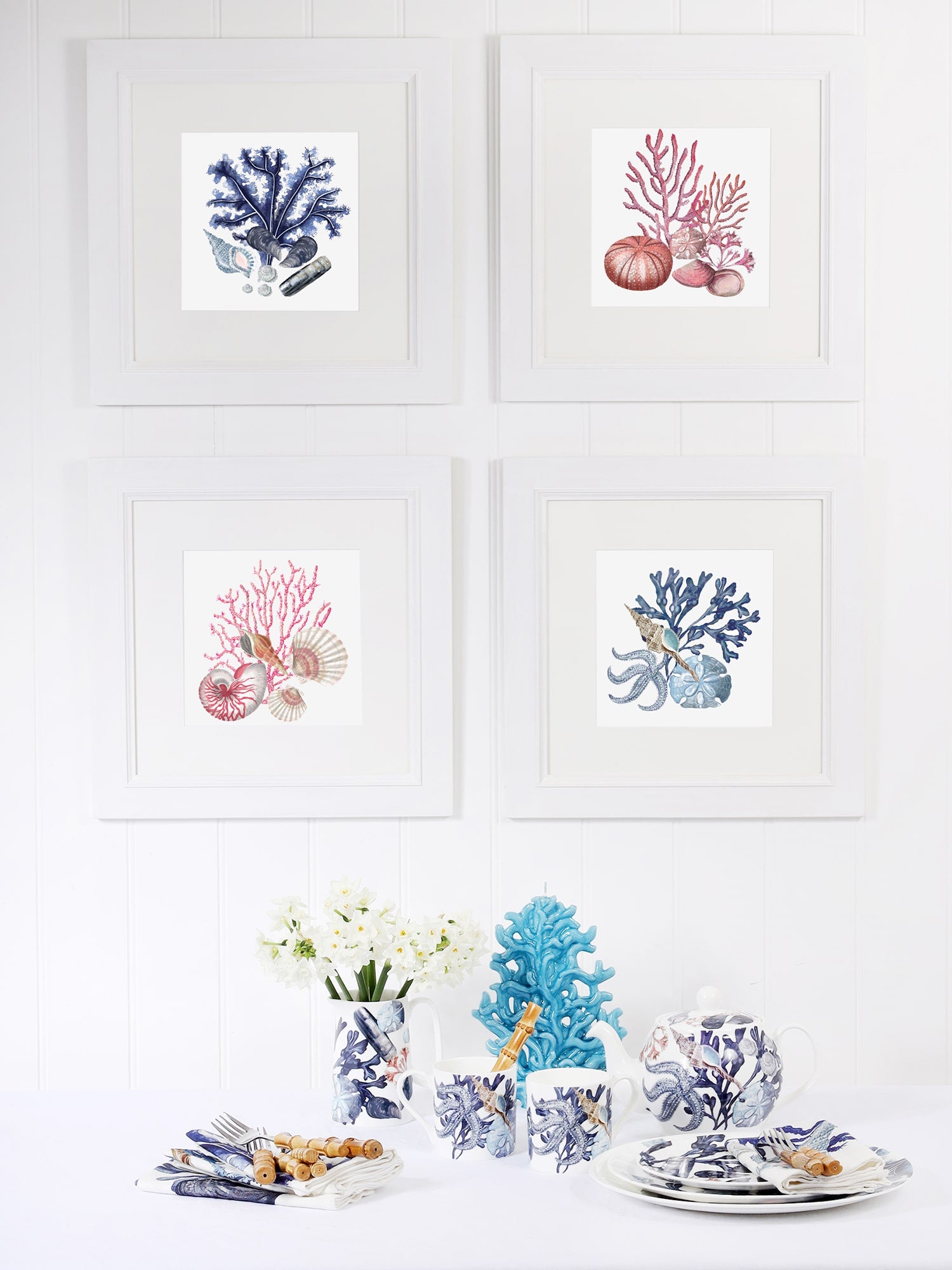 A coastal-themed dining setup with four framed coral and seashell prints in white frames on a white paneled wall. Below, a table is set with marine-inspired tableware, including mugs, plates, and a teapot featuring blue and white sea life illustrations. A vase with white flowers, a blue coral sculpture, and neatly arranged napkins with cutlery complete the ocean-inspired aesthetic.
