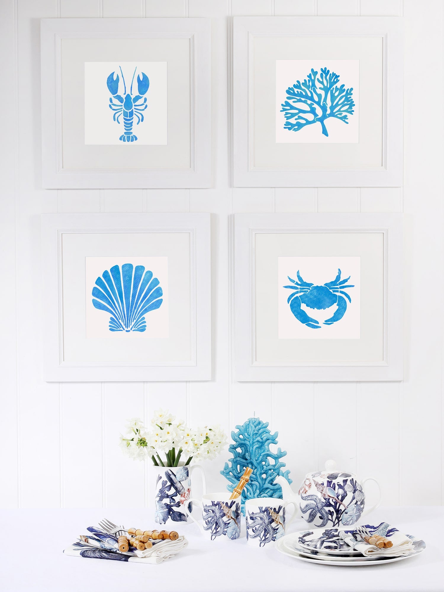 Four framed blue coastal-themed illustrations—lobster, coral, scallop shell, and crab—arranged on a white-paneled wall. Below, a table is set with ocean-inspired ceramics, white flowers, and a decorative blue coral sculpture, creating a serene coastal decor display.
