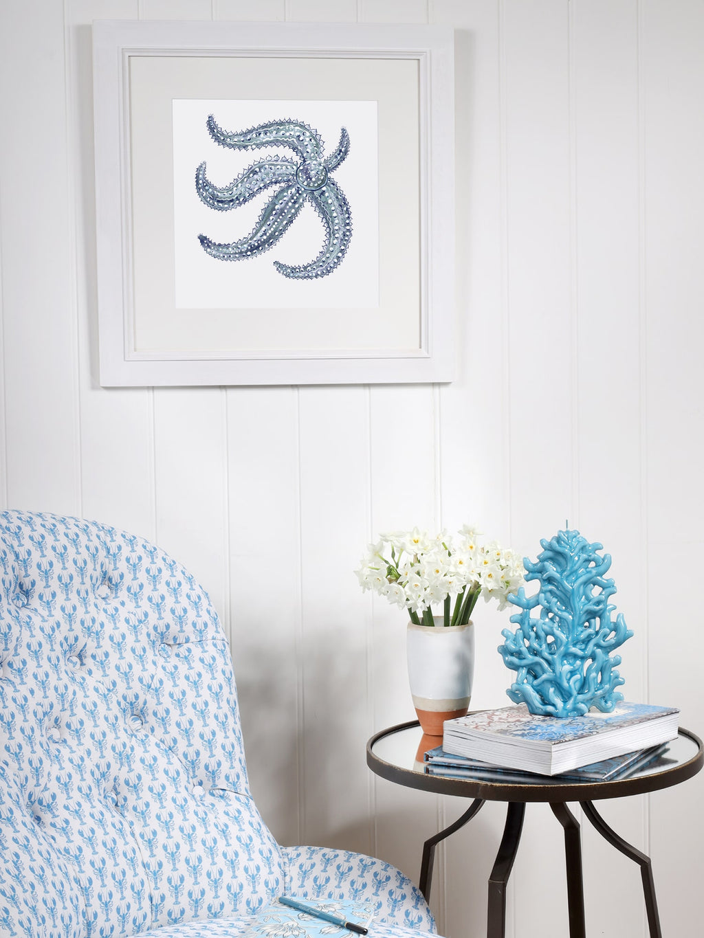 A coastal-inspired reading nook featuring a framed artwork of a detailed blue starfish on a white paneled wall. Below, a cushioned chair with blue lobster block-printed fabric sits next to a round side table. The table holds a vase with white flowers, a decorative blue coral sculpture, and a stack of books, creating a serene ocean-themed aesthetic.