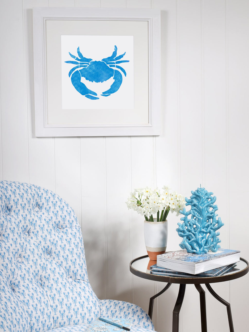 A framed blue crab illustration hangs on a white-paneled wall above a cozy corner with a blue lobster-patterned armchair. A round side table holds white flowers, books, and a decorative blue coral sculpture, creating a coastal-inspired setting.