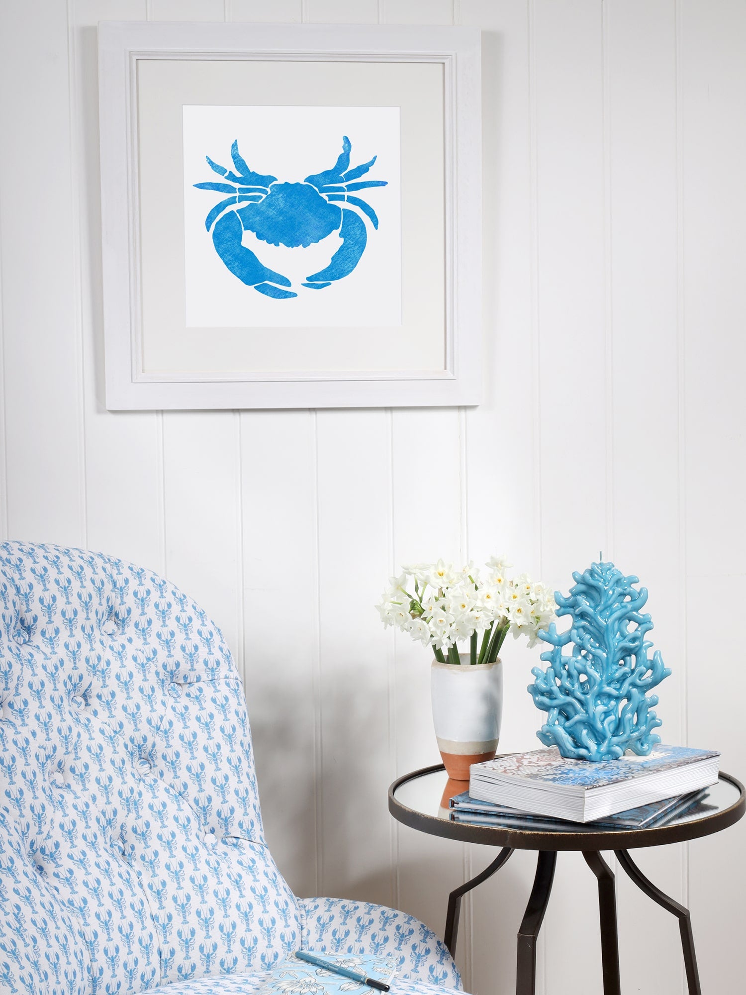 A framed blue crab illustration hangs on a white-paneled wall above a cozy corner with a blue lobster-patterned armchair. A round side table holds white flowers, books, and a decorative blue coral sculpture, creating a coastal-inspired setting.