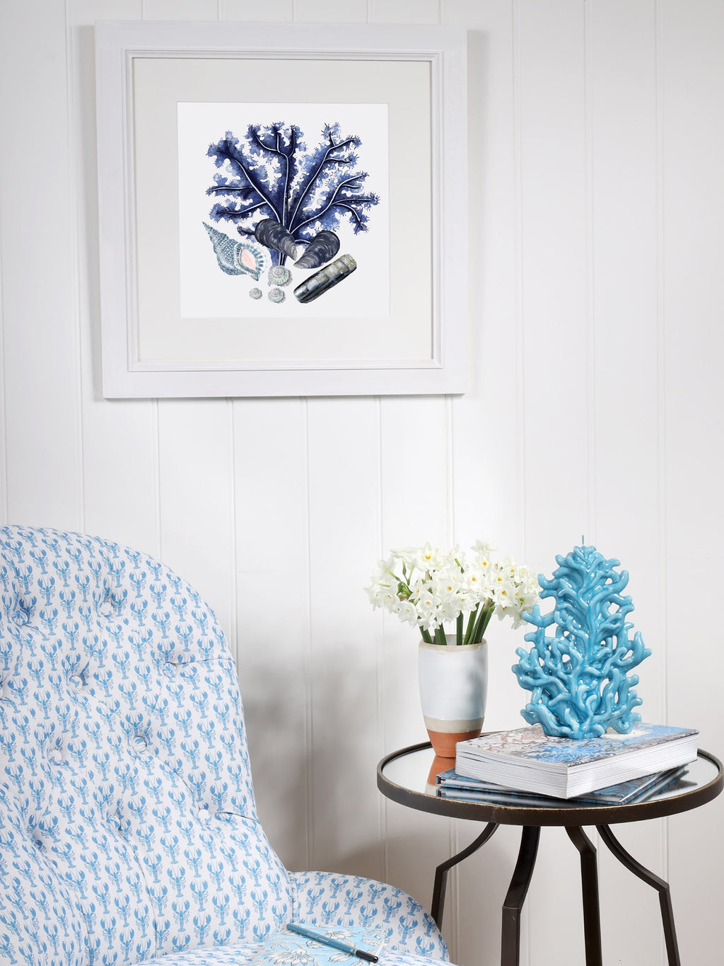 A coastal-themed corner with a framed blue coral and seashell print on a white paneled wall. Below, a cushioned chair with blue lobster block-printed fabric sits next to a round side table holding a vase of white flowers, a blue coral sculpture, and a stack of books