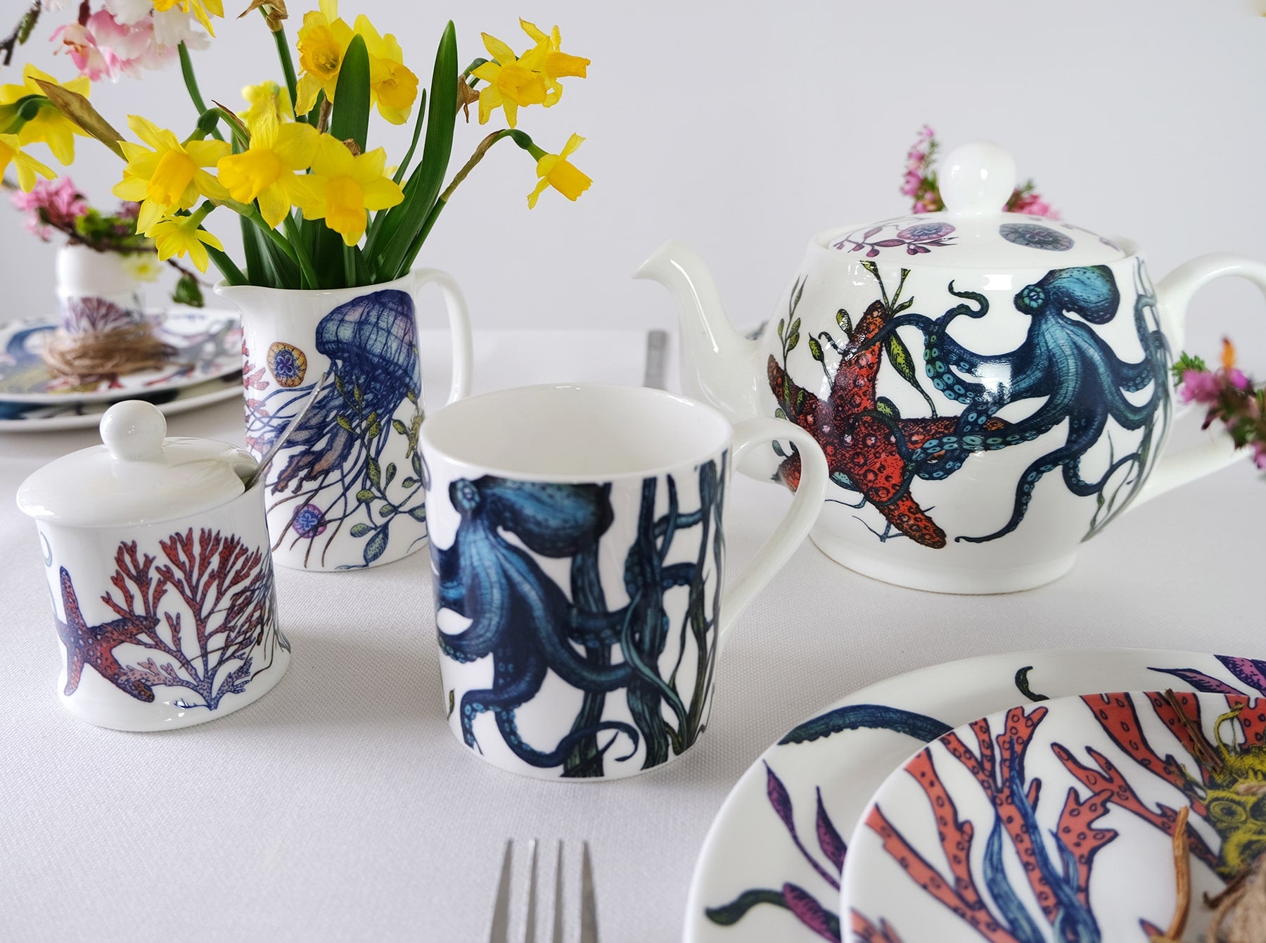 Jam Pot with lid in our bright coloured Reef range placed in front of a reef jug with Daffodils inside on a table.Next to them on the table is a Reef Teapot and various matching Reef tableware