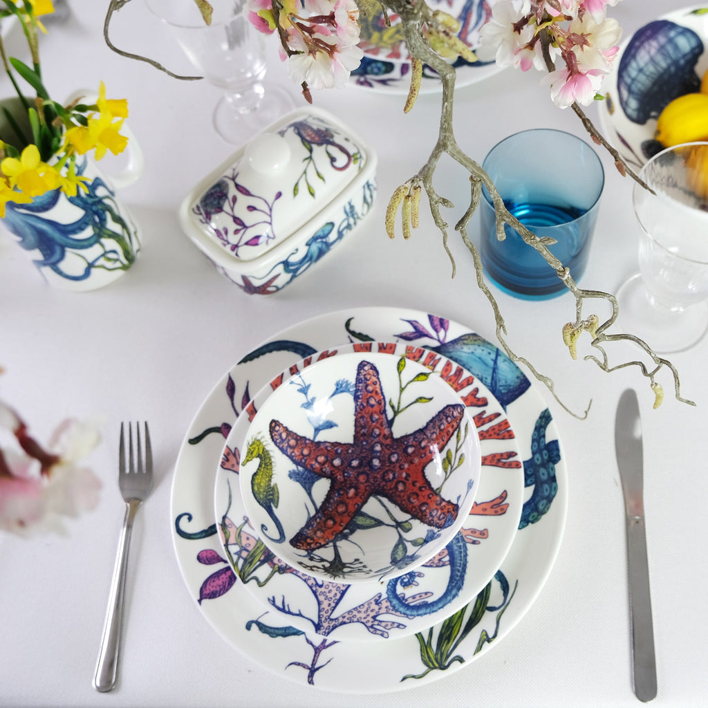 Butter dish in our brightly coloured Reef range,with Octopus,seaweed,seahorses and other sea themed designs all over the base and the lid in the centre of the table.Also on the table is a place setting in the Reef collection,pasta bowl,side and dinner plate,also you can see cutlery,coloured glasses and flowers in reef jugs