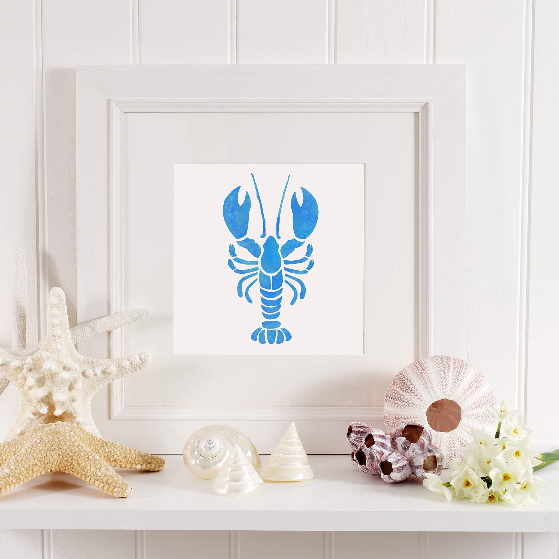 A framed blue stencil-style lobster illustration with a white mount, displayed on a coastal-themed shelf. The shelf is decorated with seashells, a starfish, a sea urchin shell, and white flowers, all set against a white paneled wall. The decor features a soft, natural color palette with blue, white, and sandy tones.