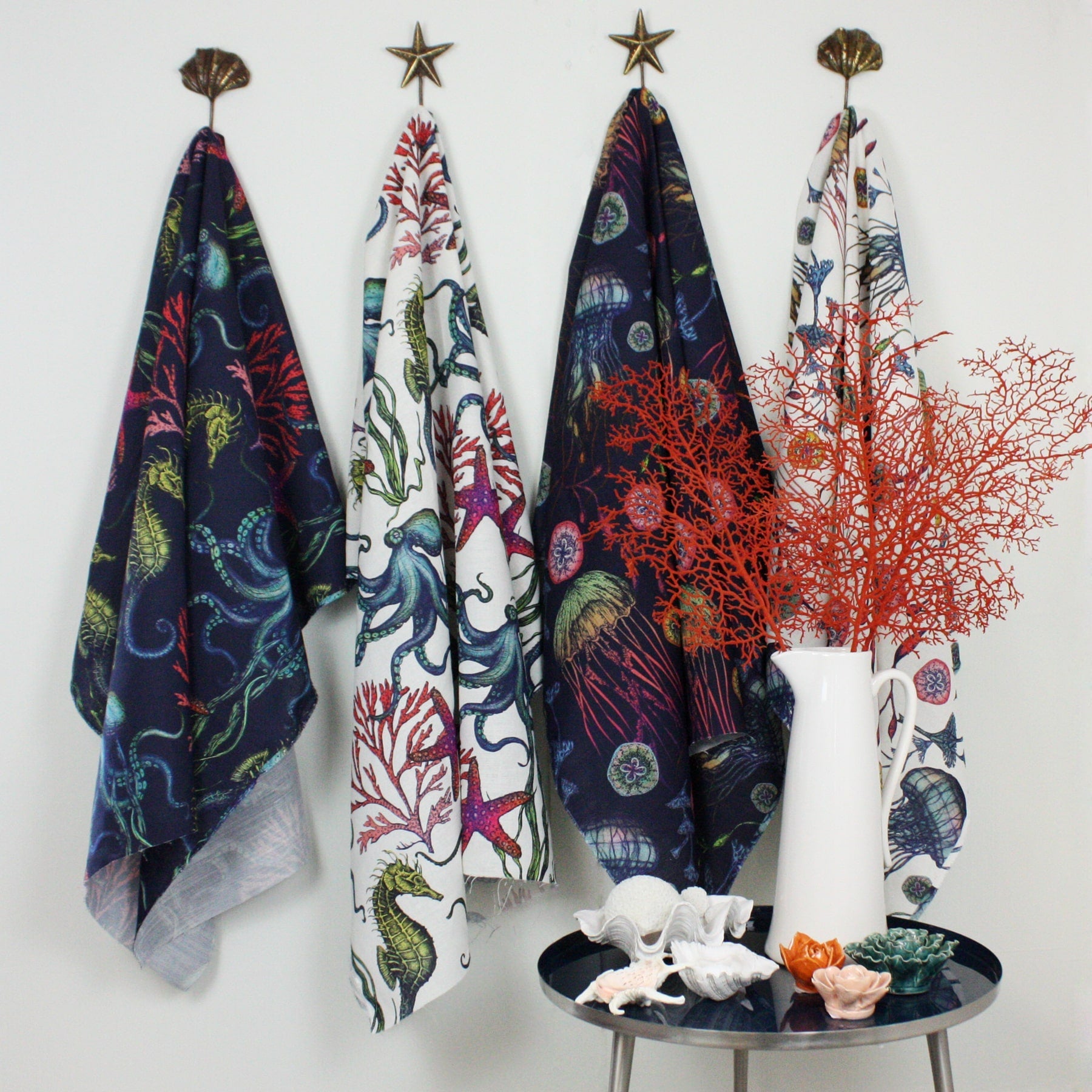 Four lengths of Cream Cornwall’s Reef fabric hanging on decorative shell and star hooks against a white wall. The fabrics feature vibrant underwater illustrations of sea creatures, coral, and jellyfish in navy and white backgrounds. Below, a round table holds ceramic sea-themed ornaments and a white jug with bright red coral branches, adding a coastal decorative touch.