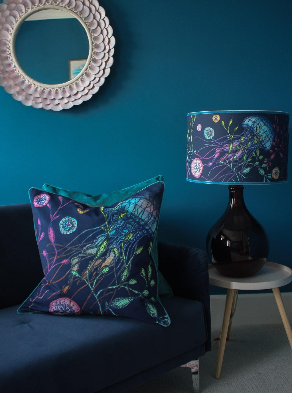 Canyons Reef Navy Shade With Jellyfish Design on a blue glass lampbase placed on a small table. Next to the table is a navy sofa with a couple of Reef cushions placed on them,behind on the Blue wall is a pink scallop shell mirror.