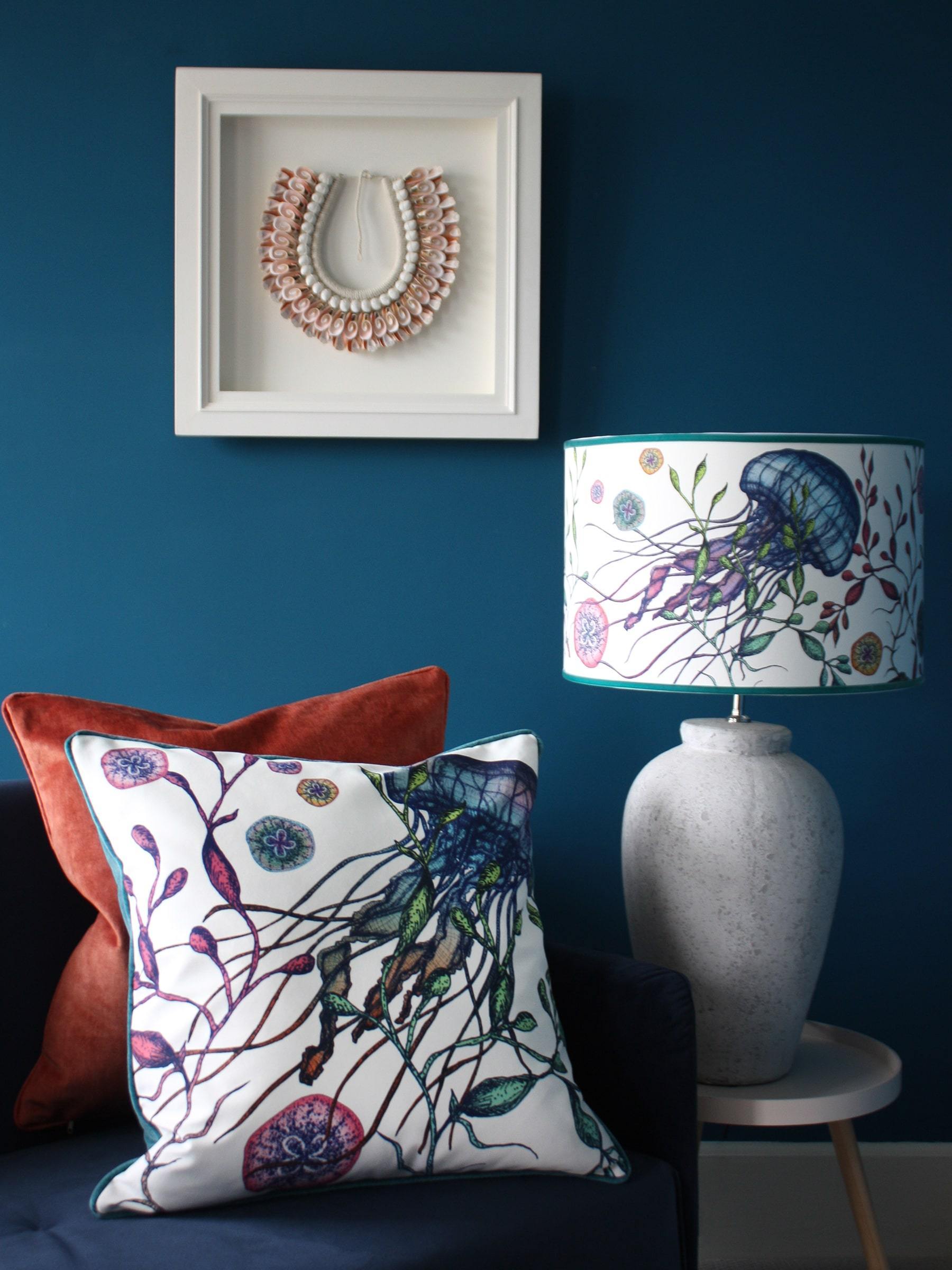 Canyons Reef White Lampshade With Jellyfish Design on a white lampbase placed on a table.Its placed next to a navy sofa with reef cushions on it,behind on a blue wall is a framed shell piece of art