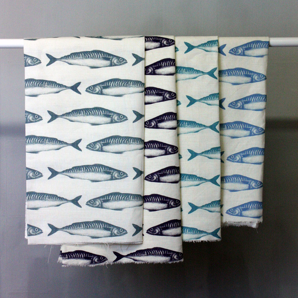 Four fabric samples hanging on a rail, each featuring a repeating mackerel fish pattern on an ecru background. The fish are illustrated in different shades of blue: dark teal, navy, turquoise, and sky blue, creating a nautical and coastal aesthetic.