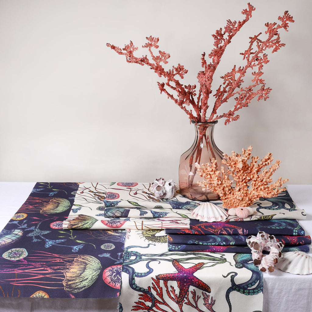 A styled table display showcasing a variety of Cream Cornwall’s Reef fabrics in both navy and off-white backgrounds, featuring vivid marine illustrations of octopuses, starfish, jellyfish, and coral. The fabrics are elegantly draped and folded beside coral-inspired decorations, seashells, and a tall pink glass vase holding faux coral branches, creating a coastal-themed aesthetic.