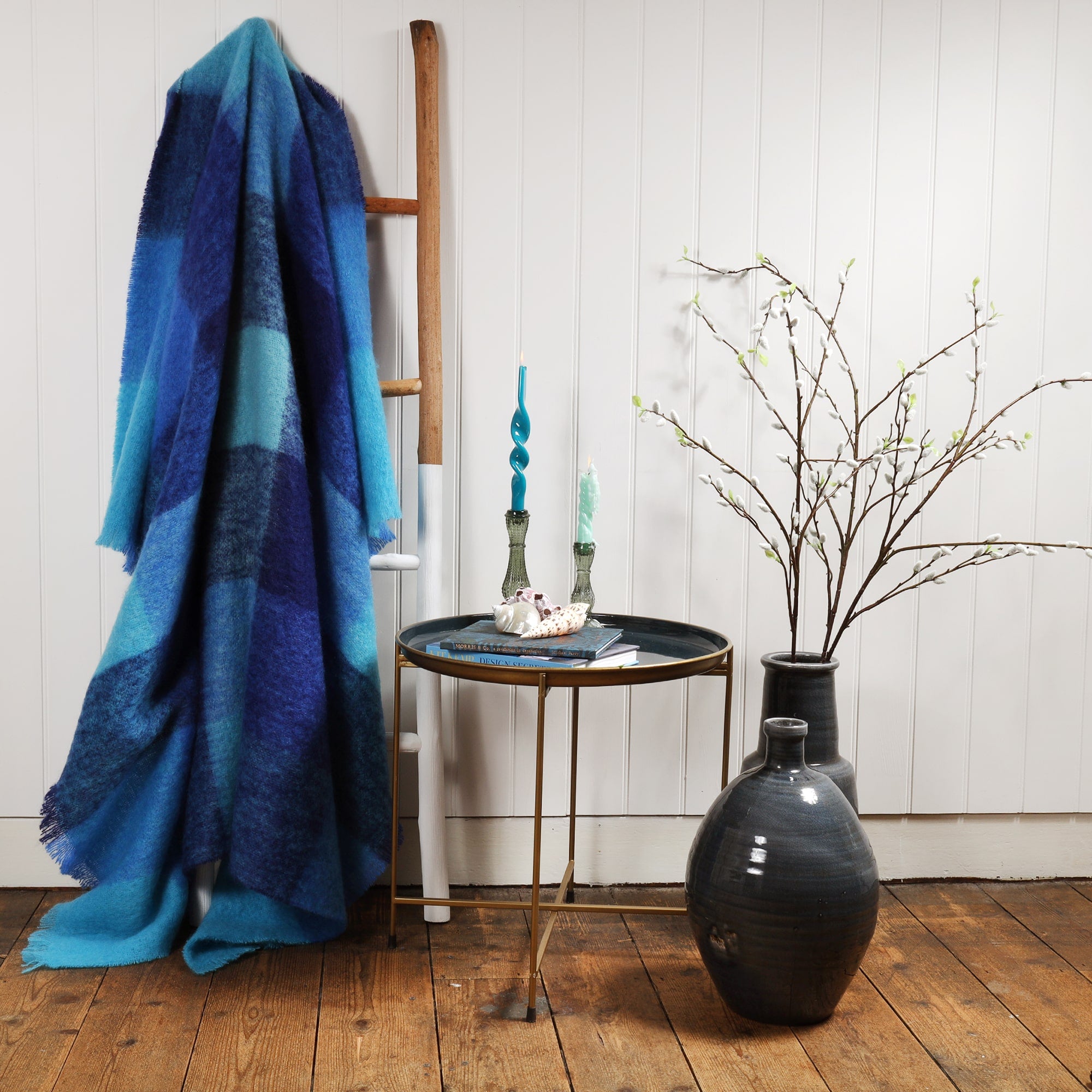 Mohair Throw in Aqua Blue check throw draped over a ladder against a wall.Next to the ladder is a table with books and a beachcomber Teapot and candles and candle holders.Next to the table are large navy vases with catkins coming out of them