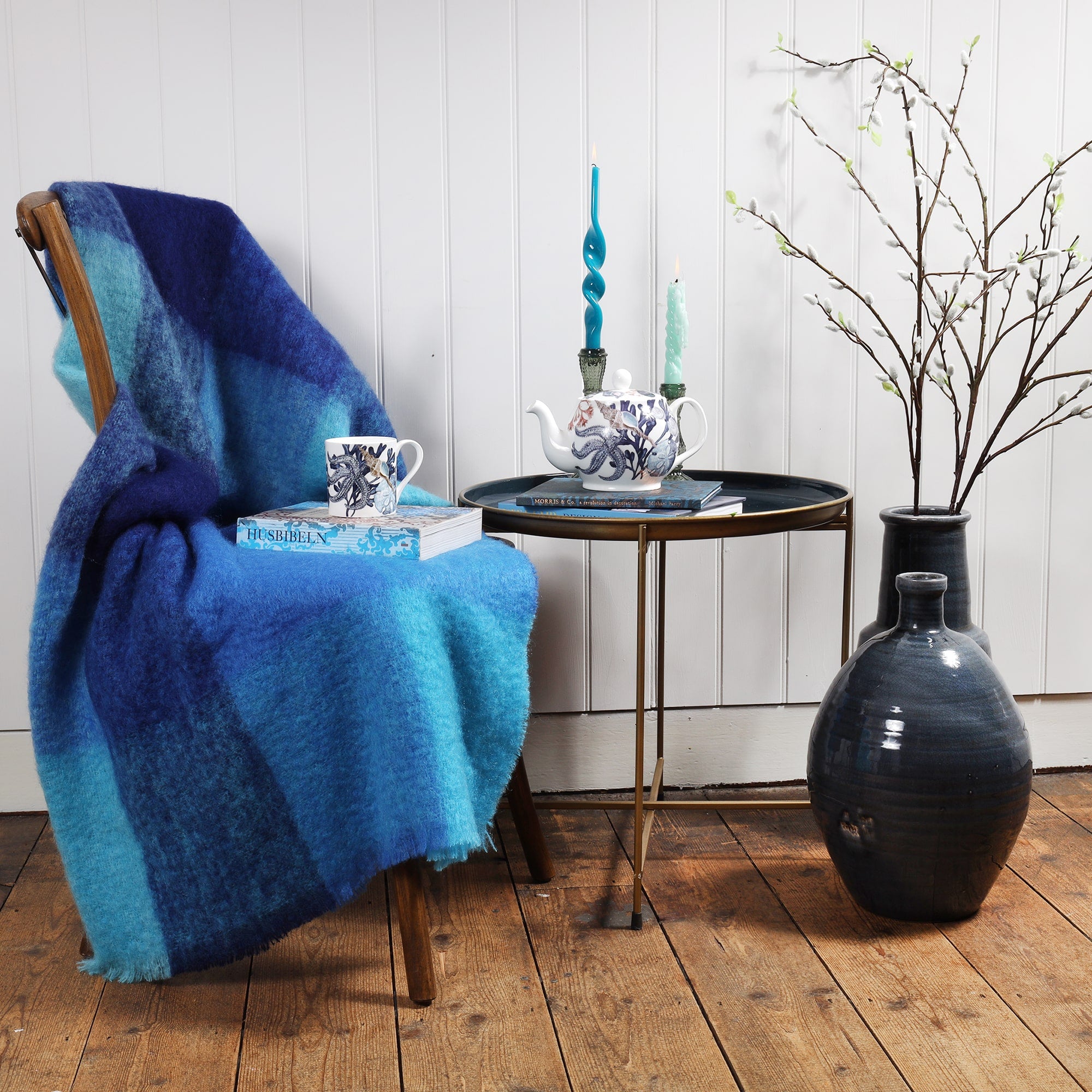 Mohair in Aqua Blue Check throw draped over a chair.Placed on the chair is a book and a mug in Beachcomber design,next to the chair is a table with books and a beachcomber Teapot and candles and candle holders.Next to the table are large navy vases with catkins coming out of the