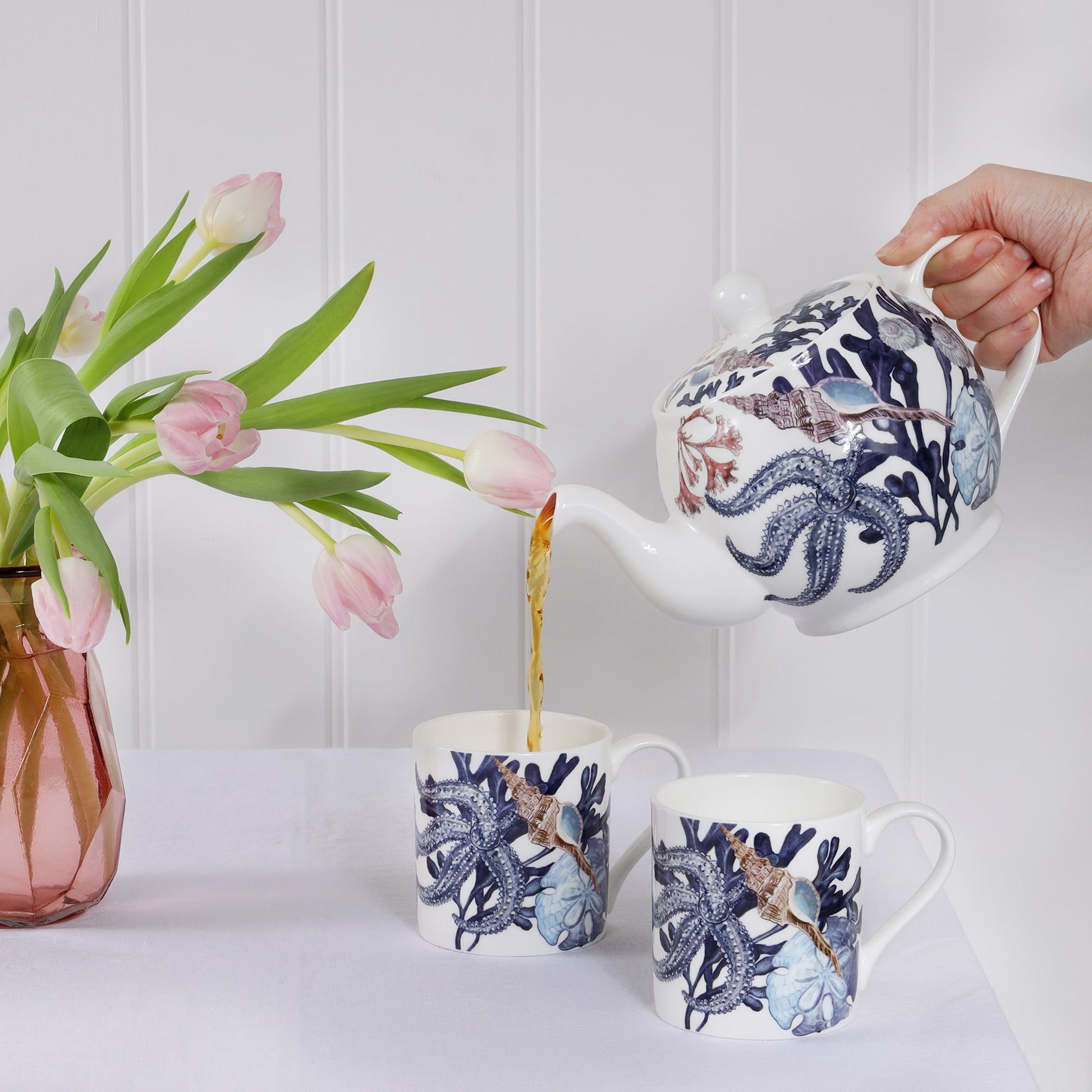 The photo shows two beachcomber mugs  on a white bench top. Tea is being poured from a beachcomber tea pot into one of the mugs. There are pink tulips in a vase to the side of the picture