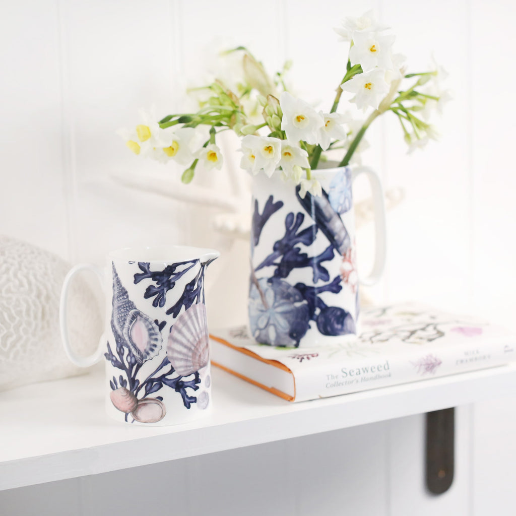 Medium size beachcomber jug next to the small beachcomber jug .In the jug are daffodils.Both jugs are on a shelf,one on a book