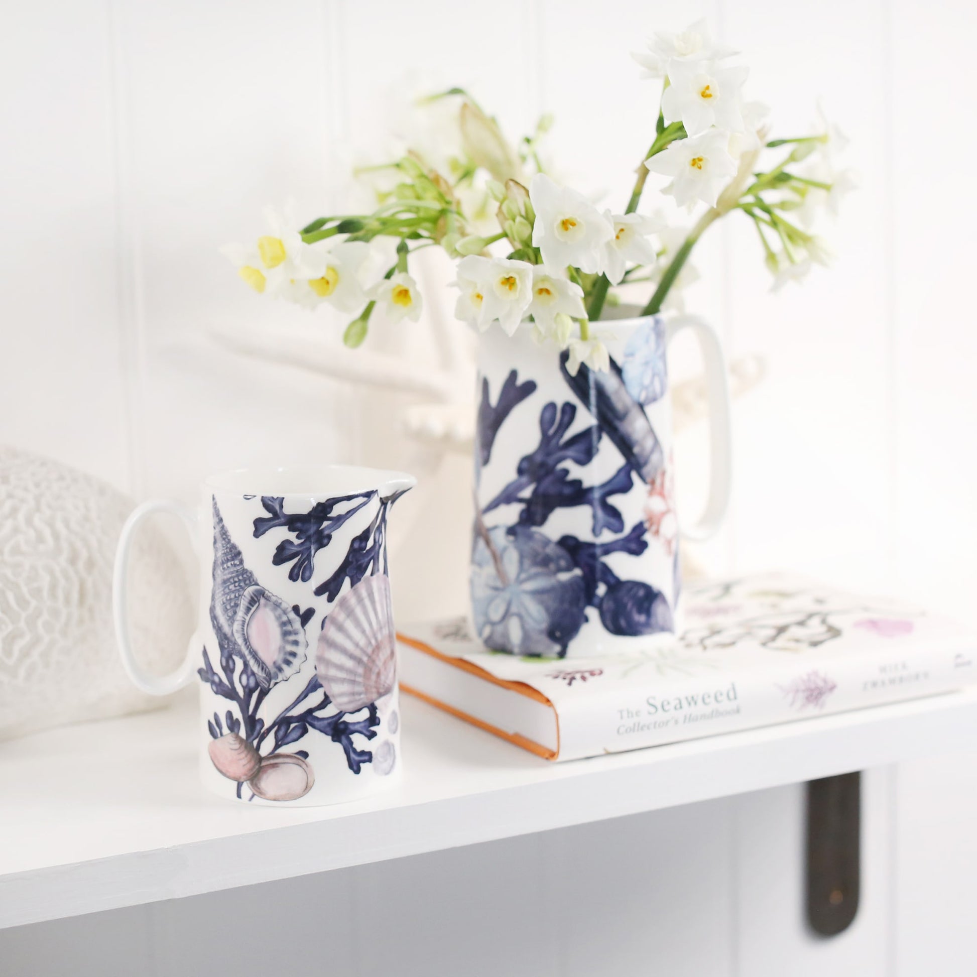Medium size beachcomber jug next to the small beachcomber jug .In the jug are daffodils.Both jugs are on a shelf,one on a book