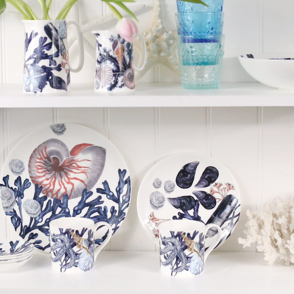 Bone china Beachcomber jugs on a shelf next to stacked blue glasses and a Beachcomber bowl.On the shelf below are Beachcomber plates on their sides,in front of those are beachcomber mugs