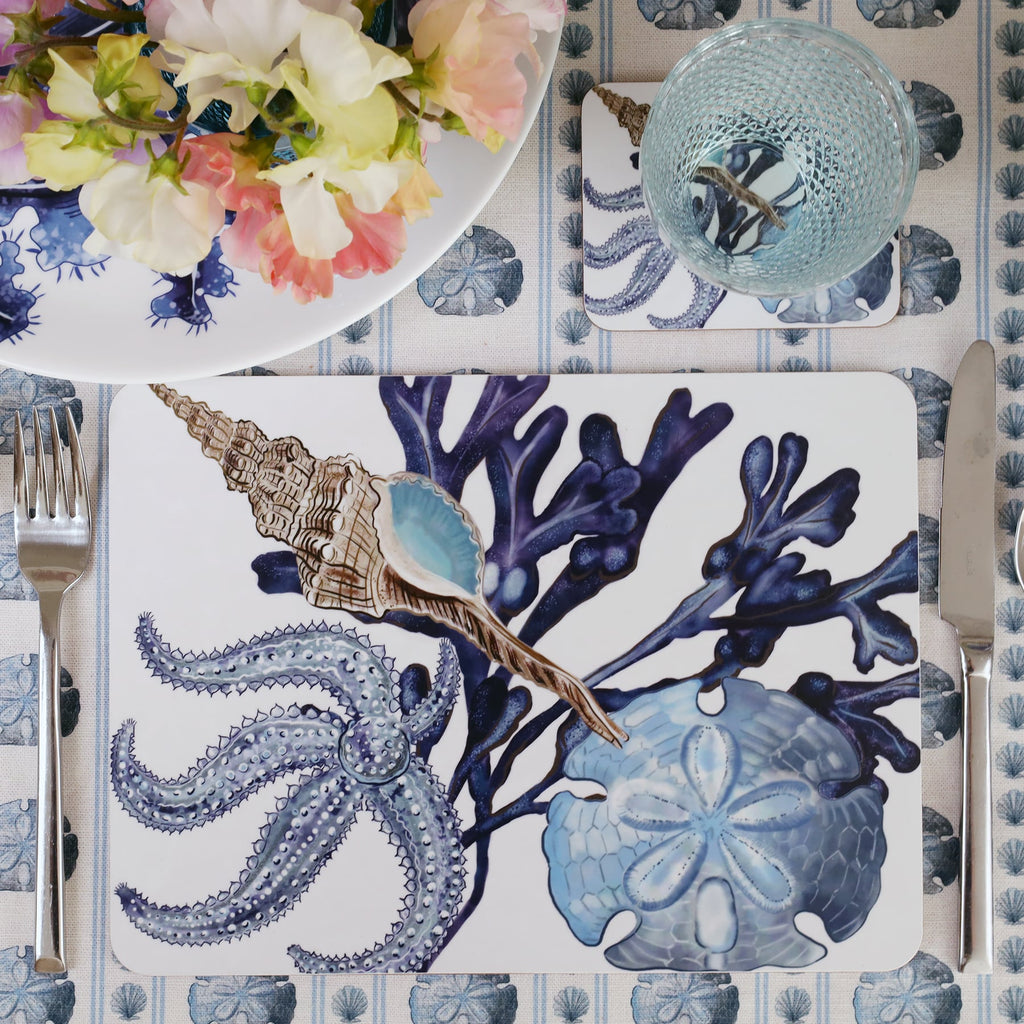 Tablecloth in the sand dollar showing a Placemat in our Beachcomber design with a matching coaster in a place setting.On the table is a bowl filled with flowers and a coloured glass on the coaster