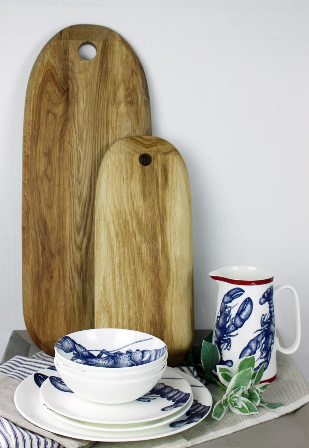Bone China White plate with hand drawn illustrations of our Classic Lobster in Navy stacked with the dinner plate,side plate and bowls and a lobster Jug.Behind them are a couple of large chopping boards lent against a wall