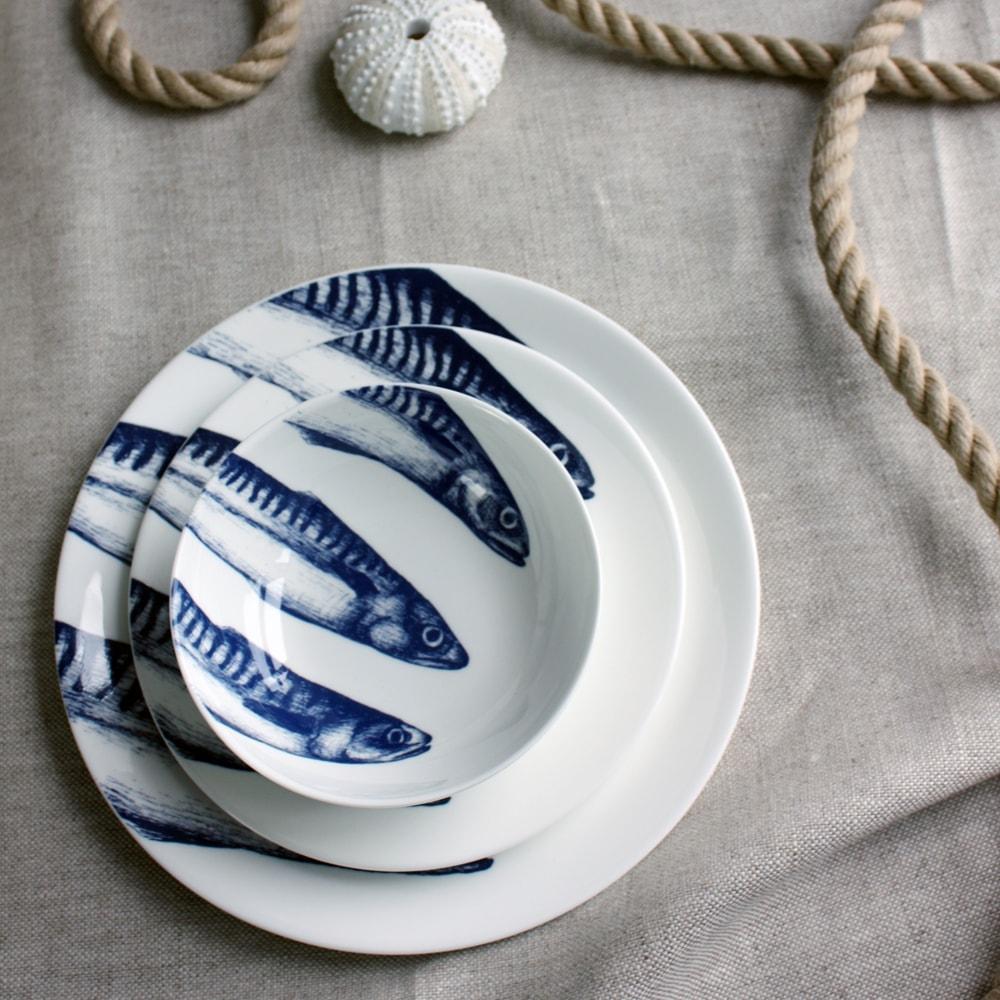 Bowl in Bone China in our Classic range in Navy and white in the Mackerel design stacked on a matching side plate and dinner plate placed on a tablecloth.On the table is a rope winding on the table and a shell
