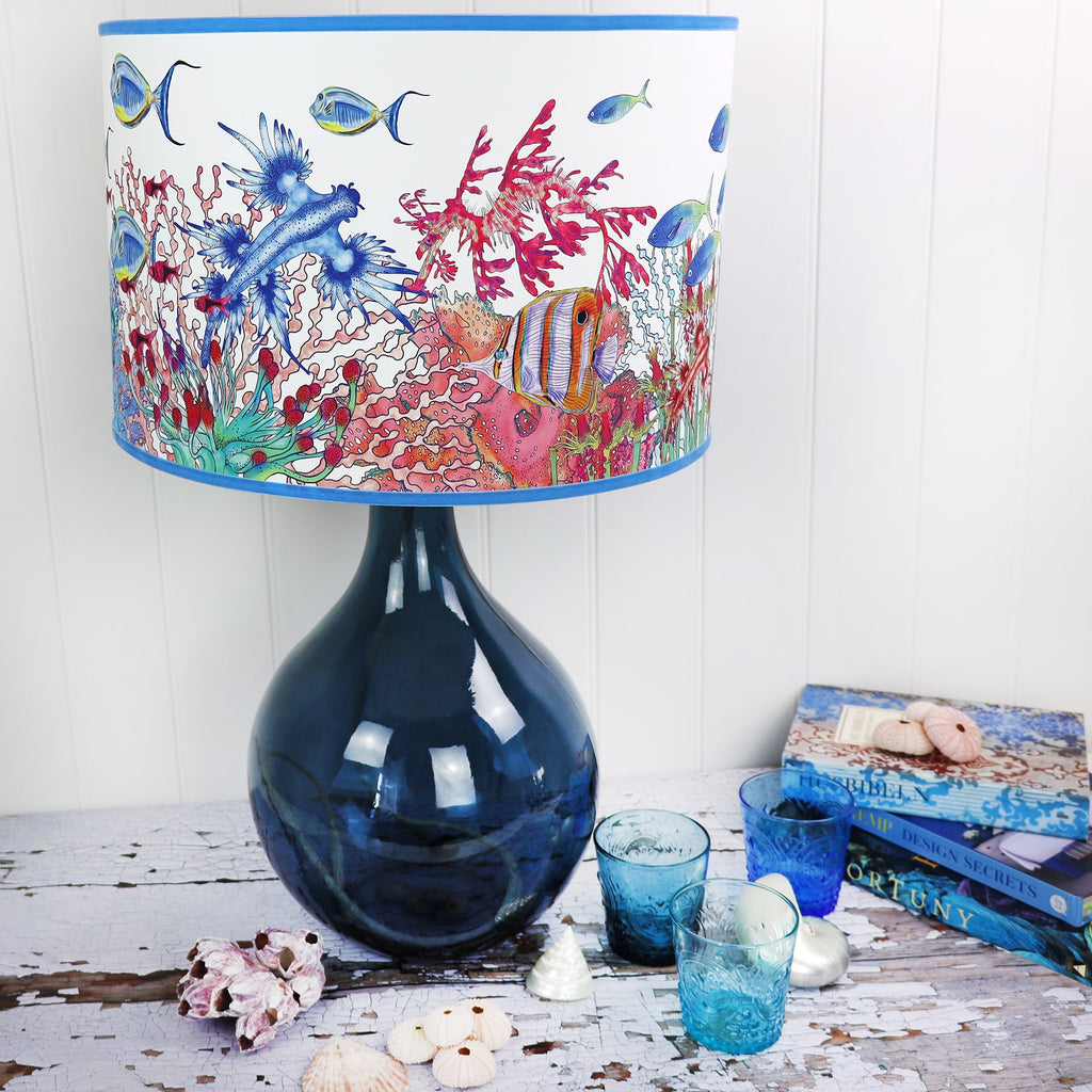 Coral Bay design lampshades in White with blue trim on a petrol glass lampbase placed on a table.Next to the lampbase is a Coral Ray plate behind a book and some shells