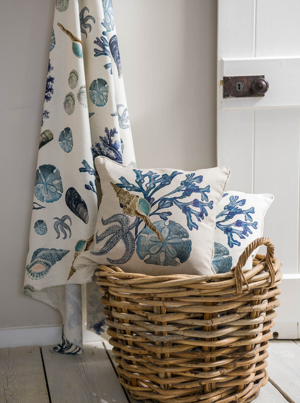 A close-up of a cozy corner featuring Cream Cornwall’s Rockpool fabric. A woven basket sits on a whitewashed wooden floor, filled with coastal-inspired cushions adorned with illustrations of seaweed, shells, starfish, and sand dollars in soft blues and neutrals. Behind the basket, matching fabric is draped casually, adding texture and a relaxed, seaside feel to the setting.