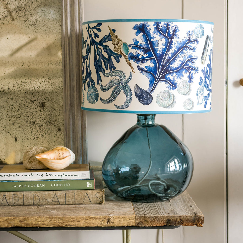 Rockpool Off White Shade With Shells,Seaweed,Sand dollars and Starfish Design in blues/browns with a blue trim on the edge of the shade placed on a glass lampbase.Placed on a wooden table next to some books.