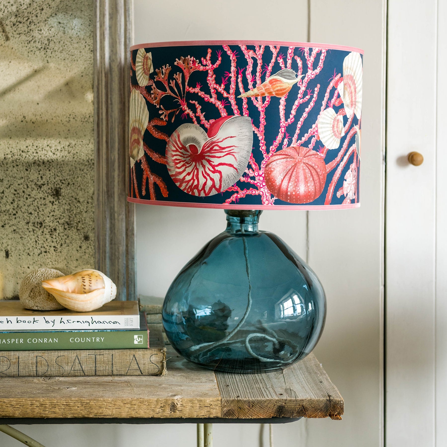 Shell Seeker Ink Blue Shade With Shells,Seaweed,Sea Urchin and Whelks Design in pinks/whites with a pink trim on the edge of the shade.Placed on a petrol blue glass lampbase on a table next to a stack of books