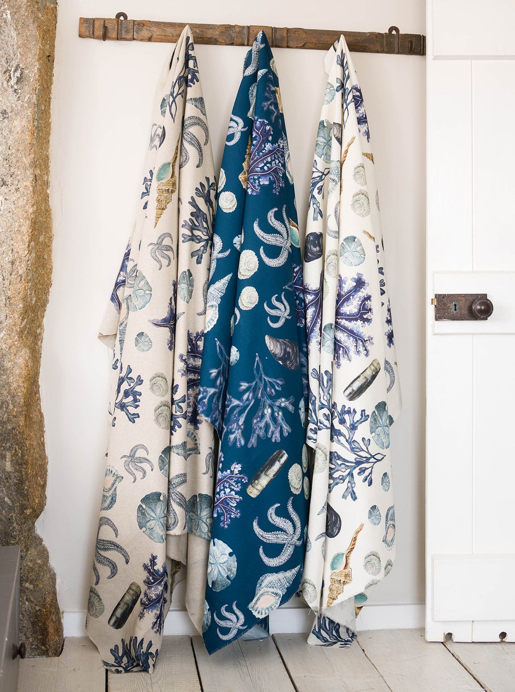 Three lengths of Cream Cornwall’s Rockpool fabric designs are draped from a rustic wooden peg rail against a white wall. The fabric features intricate marine illustrations of shells, sea urchins, corals, and starfish in soft coastal blues and sandy neutrals, with the centre fabric on a rich teal background. The natural textures and sea-inspired patterns evoke a relaxed, coastal home aesthetic.