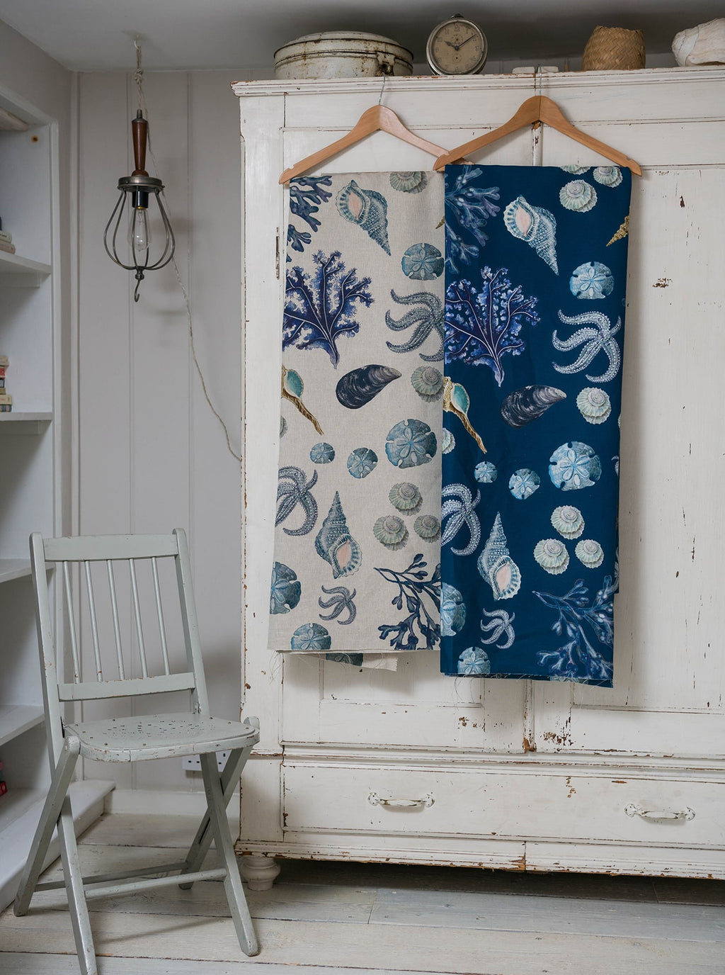 Two lengths of Cream Cornwall's Rockpool fabric hang side by side on wooden hangers against a vintage white wardrobe. One fabric has a natural linen background, the other a deep teal. Both feature detailed coastal illustrations of shells, seaweed, starfish, and sand dollars in soft blues and neutrals. A distressed pale green wooden chair sits nearby in the rustic room.