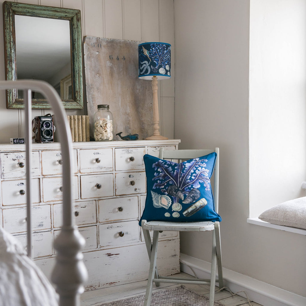 Rockpool Navy Shade With Shells,Seaweed,Sand dollars and Starfish Design in blues/browns with a blue trim on the edge of the shade on a wooden lampbase.This is placed on a chest of drawers with a camers,books and other items.In front of the chest is a chair with a Rockpool cushion.