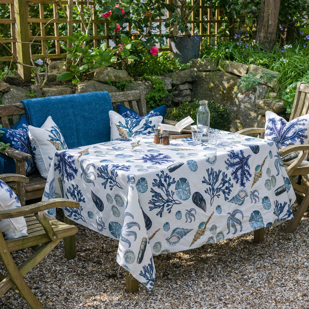 An inviting garden seating area with a wooden table and chairs set on a gravel surface. The table is draped with a Cream Cornwall Rockpool fabric tablecloth, decorated with blue and neutral sea-themed illustrations of coral, shells, and starfish. Matching scatter cushions with coastal designs adorn the surrounding chairs and bench. A glass bottle, drinking glasses, an open book, and condiments are placed on the table. Lush greenery, flowering plants, and a stone wall complete the tranquil outdoor setting.