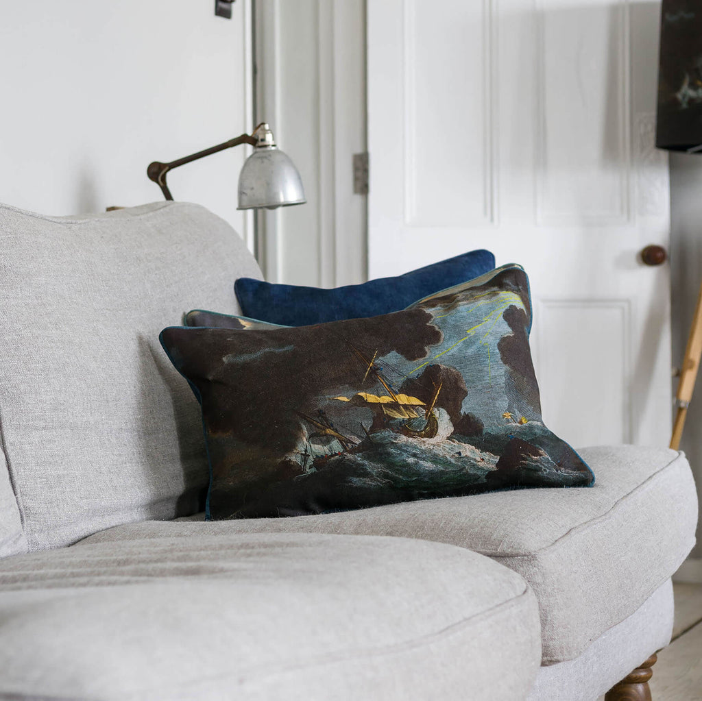 Shipwreck Night Rectangle Cushion Cover showing a couple of ships in heavy stormy,thundery weather placed on a sofa with other cushions.You can see the door behind and a Tripod in the background.