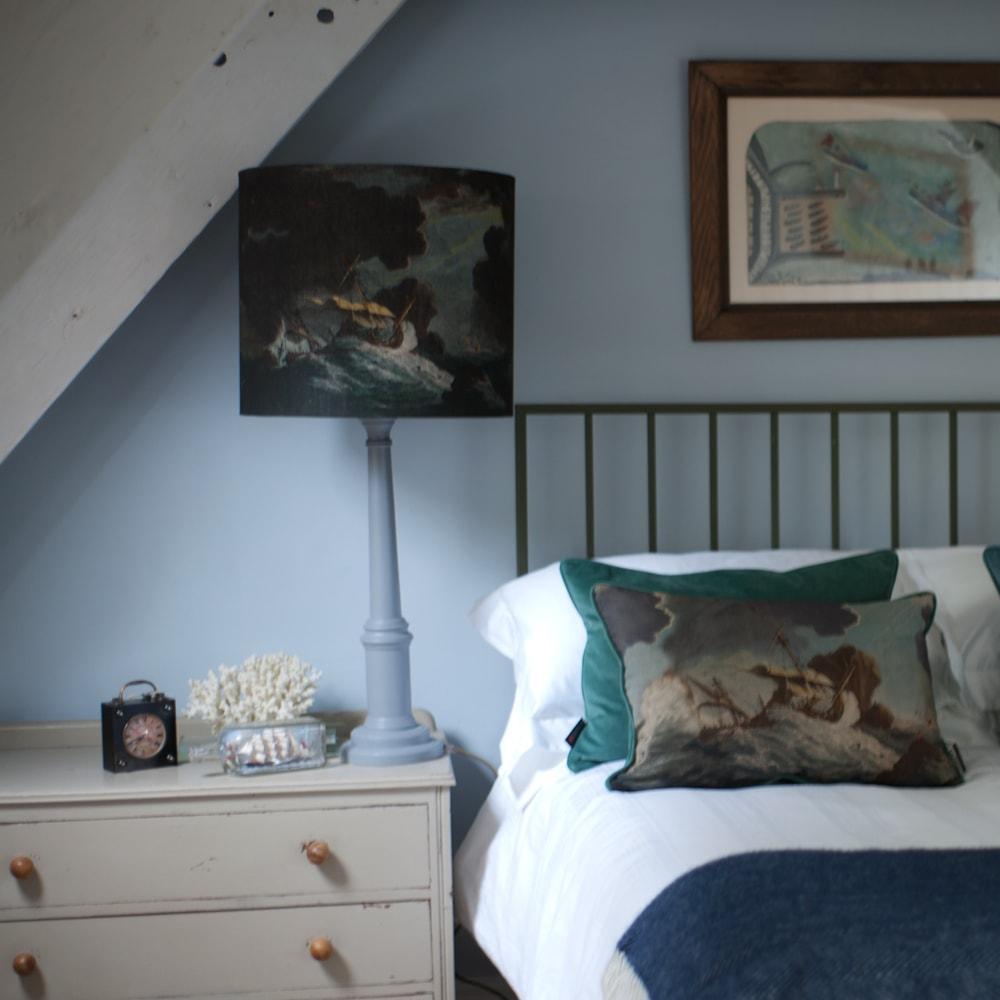 Shipwreck Day Rectangle Cushion placed on a navy throw on a bed.Next to the bed is a sideboard with a lampbase and a matching shipwreck lampshade on the base.Behind the bed is an Alfred Wallis style painting