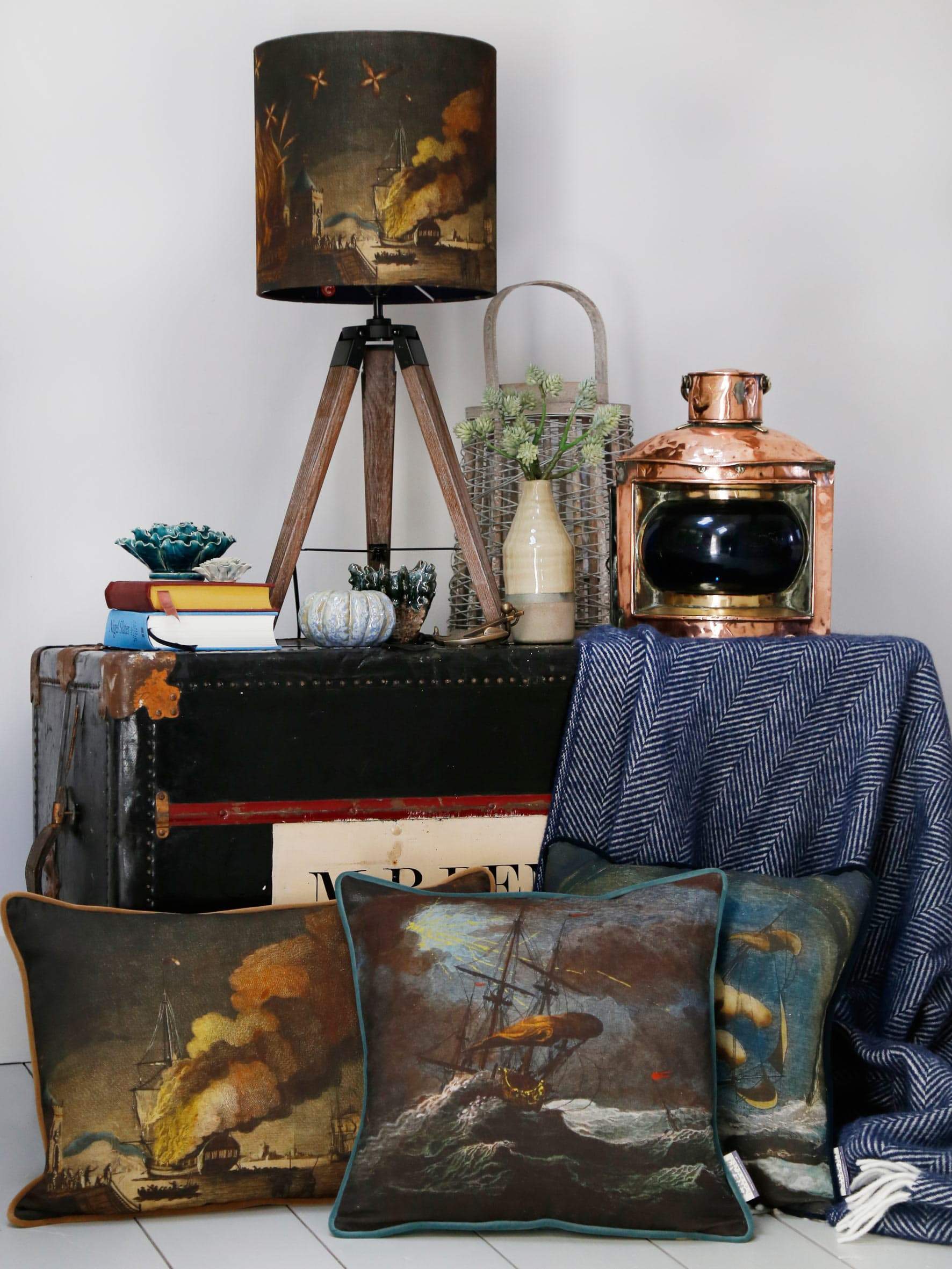 Shipwreck Night Cushion Square Cover with other shipwreck cushions placed in front of an old style trunk.On the trunk is a tripod with a shipwreck lampshade,candle holder,books and a brass ships lantern
