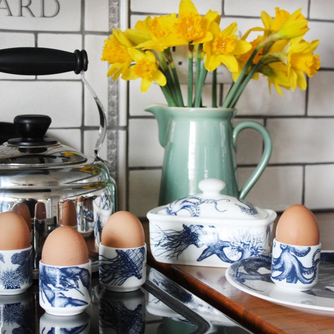 KItchen scene,showing a worktop with a jug with daffodils in the background,a classic design butterdish and several egg cups holding boiled eggs in the Jellyfish,Octopus,Seahorse and Lobster design placed in front of a kettle