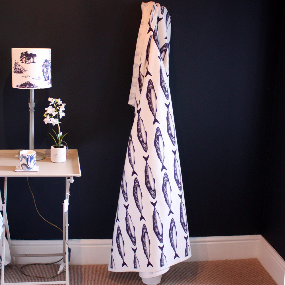 A piece of fabric with a bold navy mackerel fish print on an ecru background is draped in the corner of a room with dark blue walls. To the left, a small table holds a white lampshade with a nautical toile design, a potted white orchid, and a mug, creating a coastal-themed interior display.